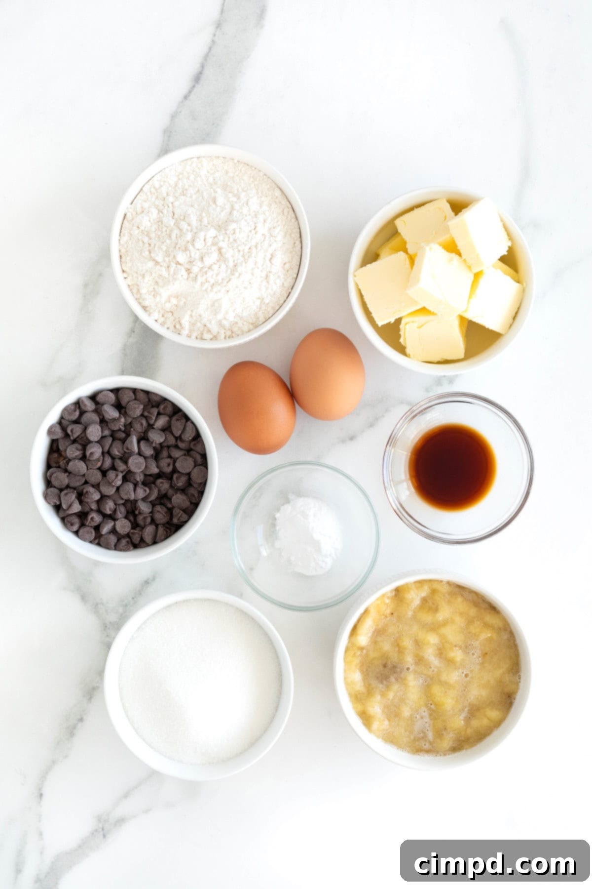 Various ingredients for chocolate chip banana bread laid out in small white and glass bowls on a pristine white marble countertop, including melted butter, sugar, flour, eggs, and chocolate chips.
