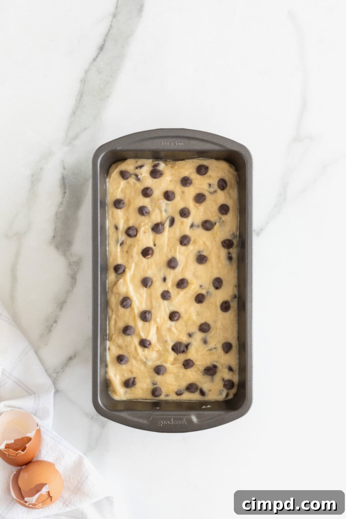 Smooth chocolate chip banana bread batter poured evenly into a silver aluminum loaf pan, ready for baking on a white marble countertop.
