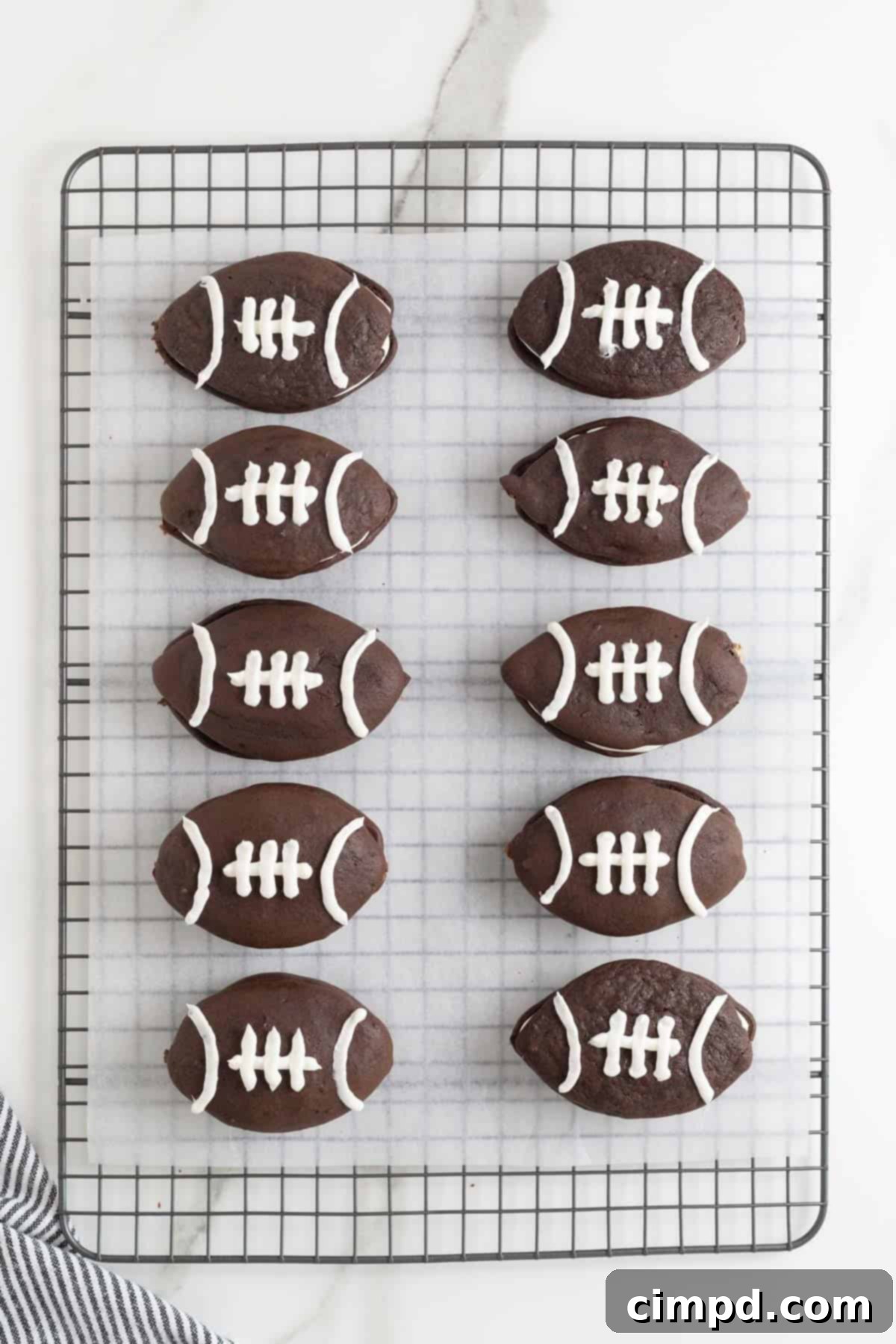 10 football shaped chocolate whoopie pies on a parchment lined cooling rack.