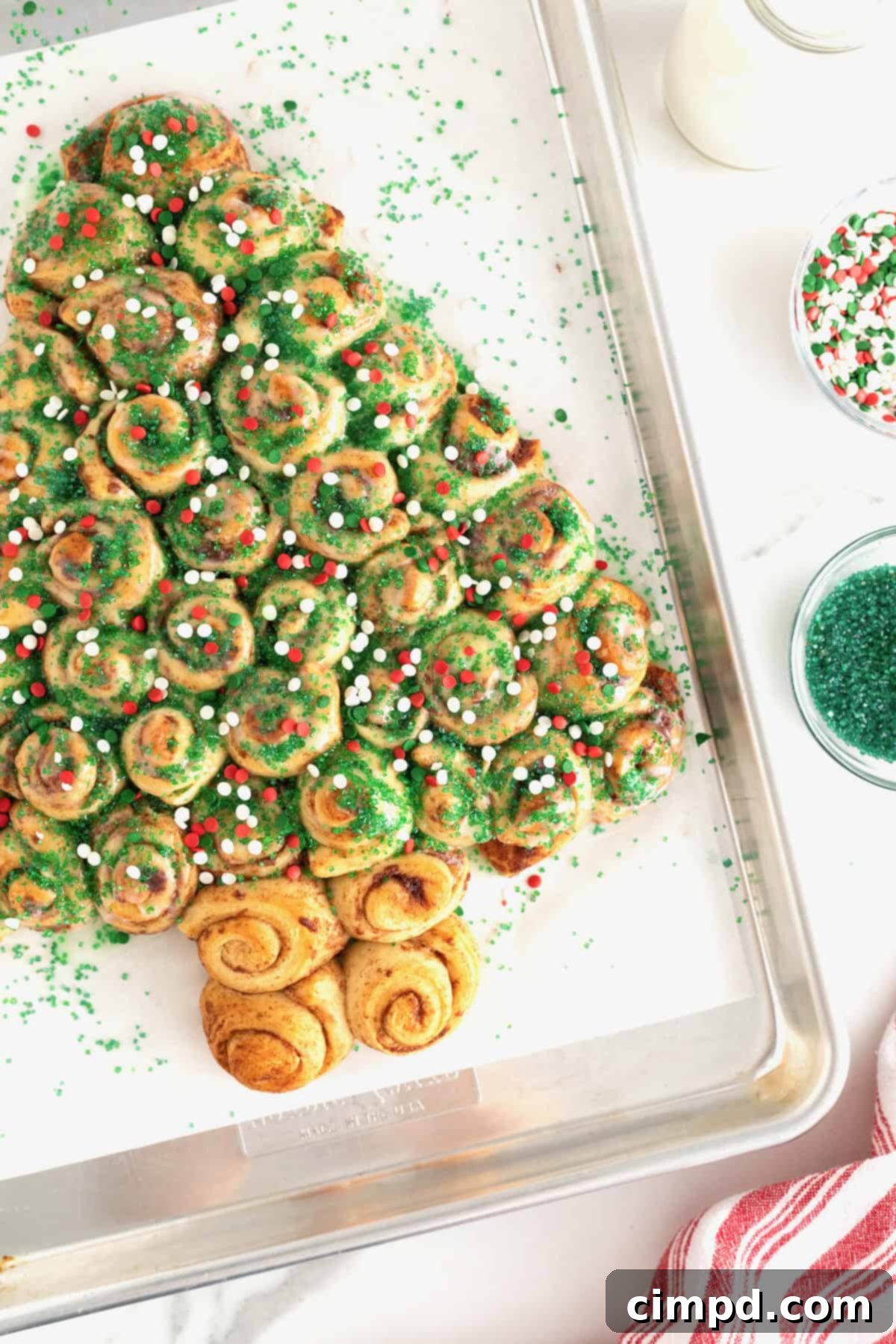 A festive cinnamon roll Christmas tree, beautifully decorated with green sugar crystals and red and green holiday sprinkles, presented on a parchment-lined baking sheet, emphasizing its delicious appeal.