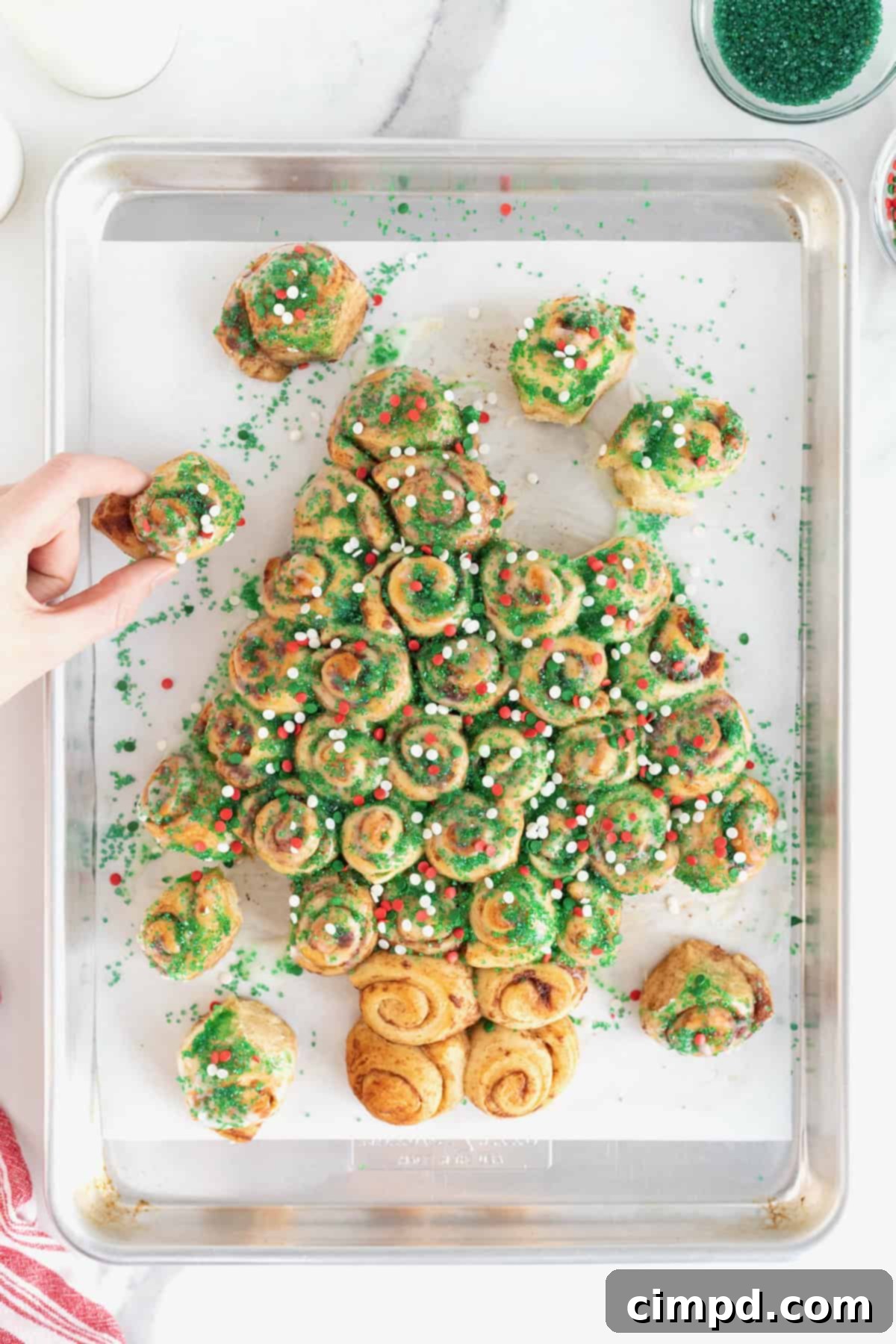 A hand reaching to pull a piece from a festive Christmas tree-shaped cinnamon roll pull-apart, adorned with colorful holiday sprinkles.