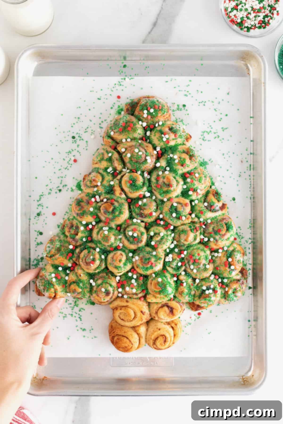 A hand reaching to pull a piece from a festive Christmas tree-shaped cinnamon roll pull-apart, adorned with colorful holiday sprinkles.