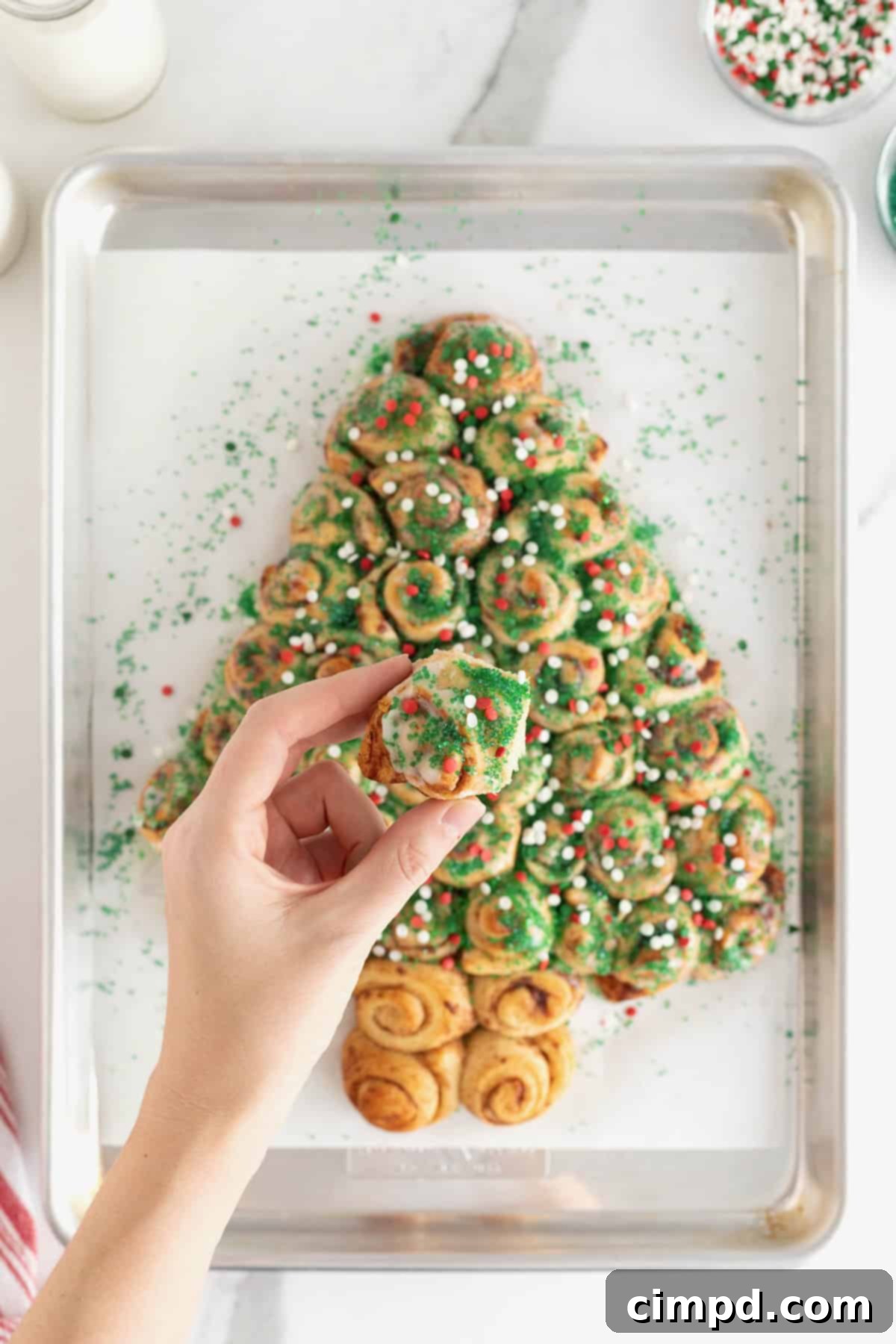 A hand reaching to pull a piece from a festive Christmas tree-shaped cinnamon roll pull-apart, adorned with colorful holiday sprinkles.