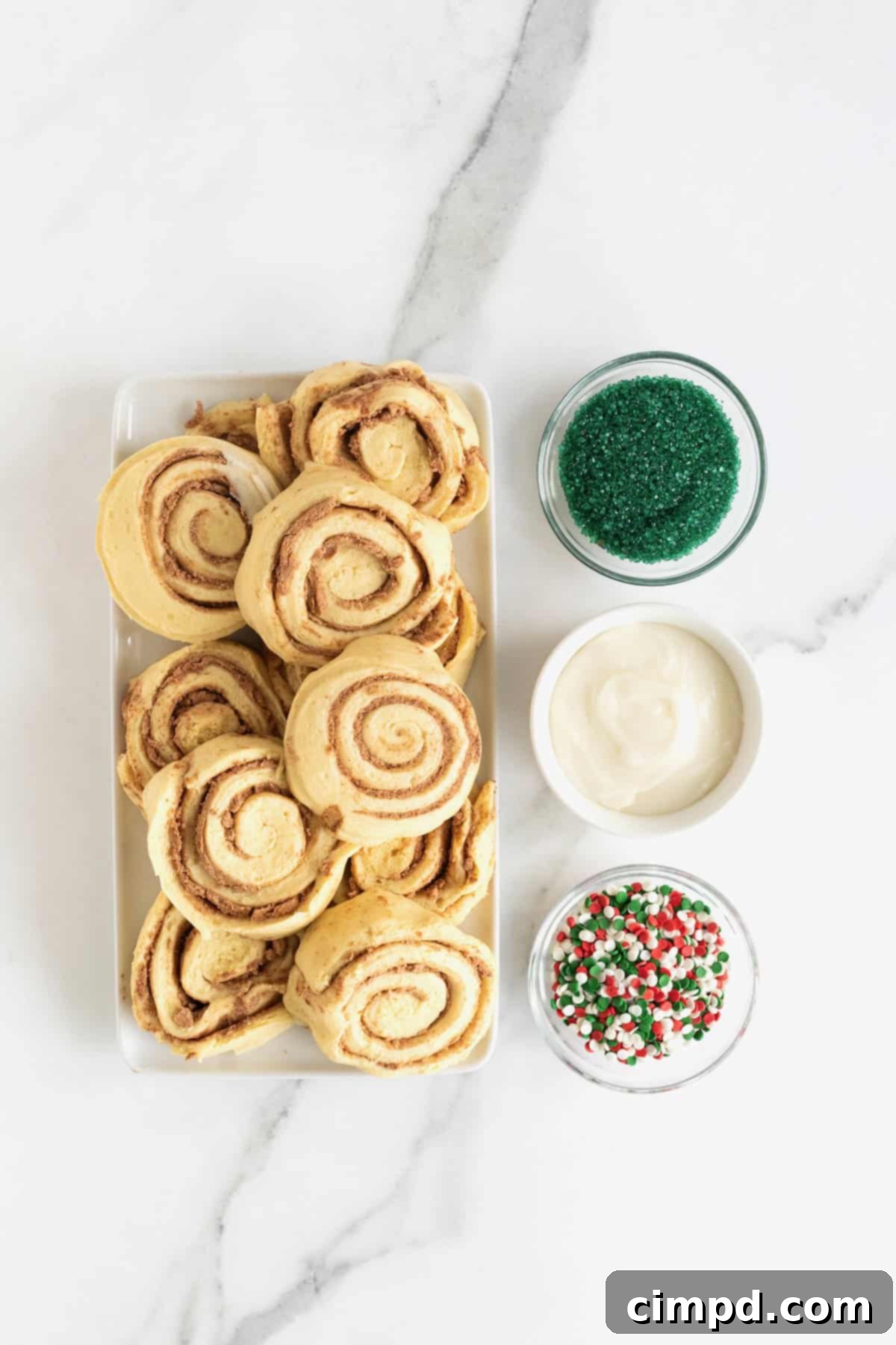 Ingredients to make a Christmas tree cinnamon roll in white dishes on a white marble counter, including cinnamon roll cans, green sugar crystals, and holiday sprinkles.