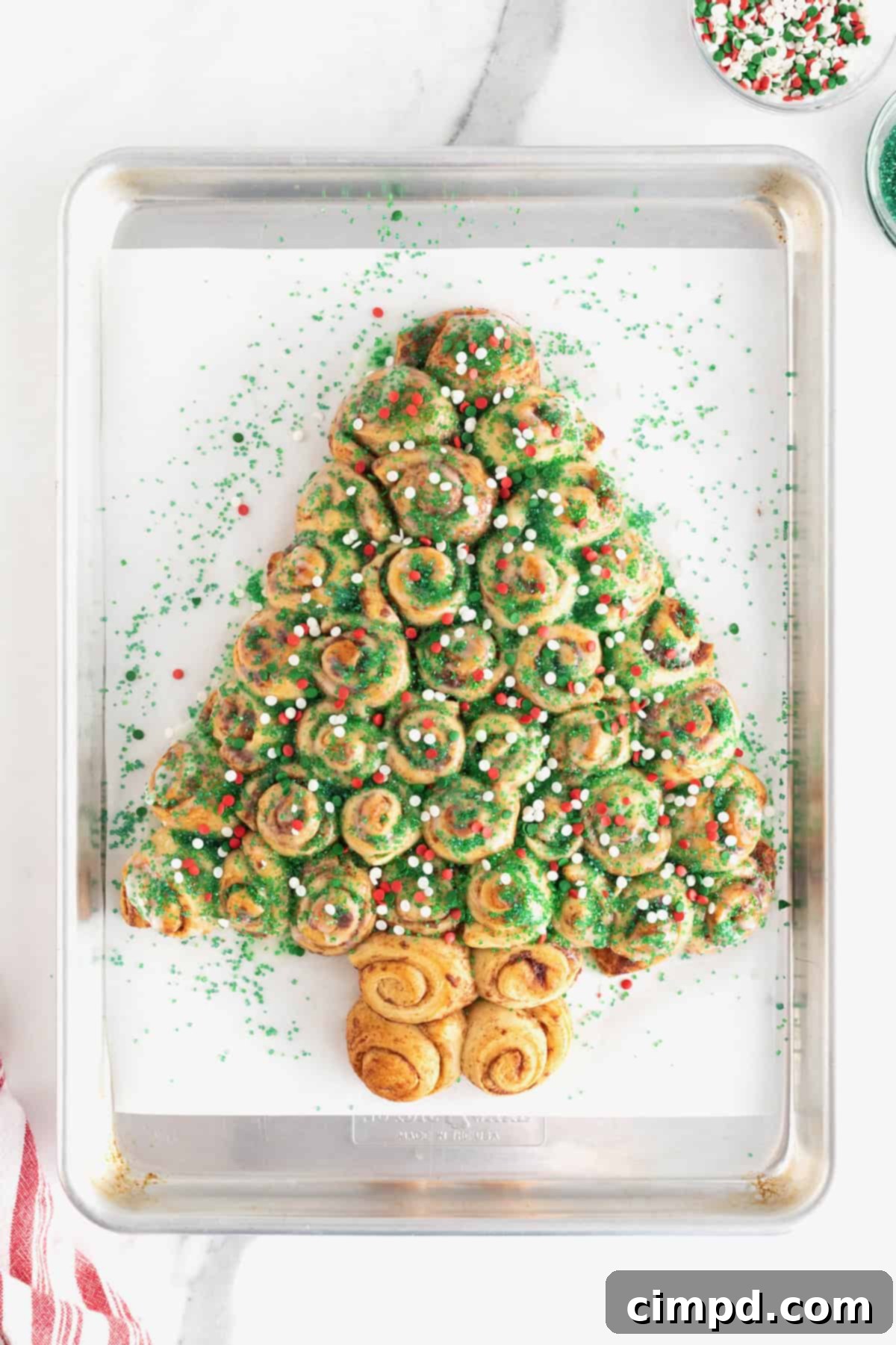 A beautifully decorated cinnamon roll Christmas tree, adorned with green sugar crystals and festive red and green holiday sprinkles, presented on a parchment-lined baking sheet.