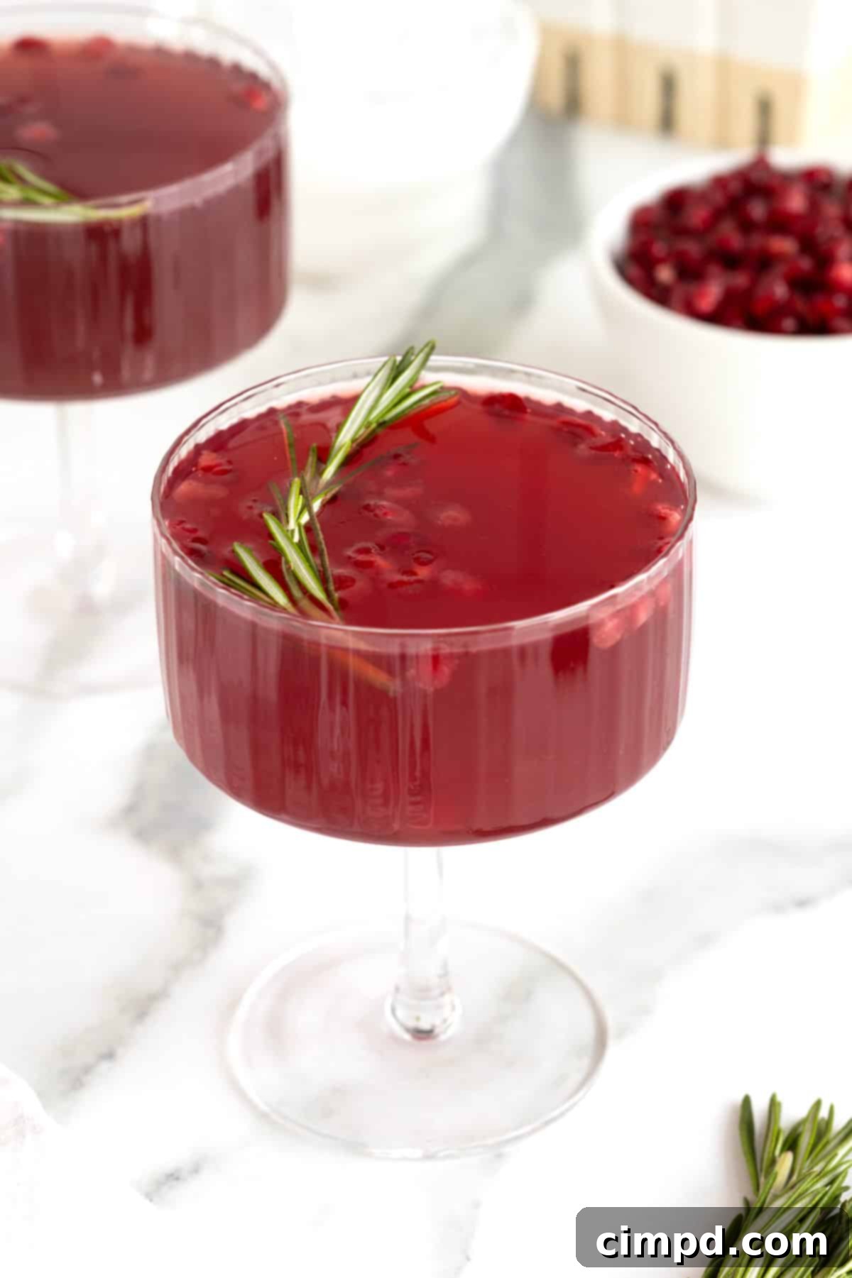 A vibrant, dark red pomegranate rosemary cocktail in an elegant stemmed glass, garnished with fresh pomegranate seeds and a sprig of aromatic rosemary.