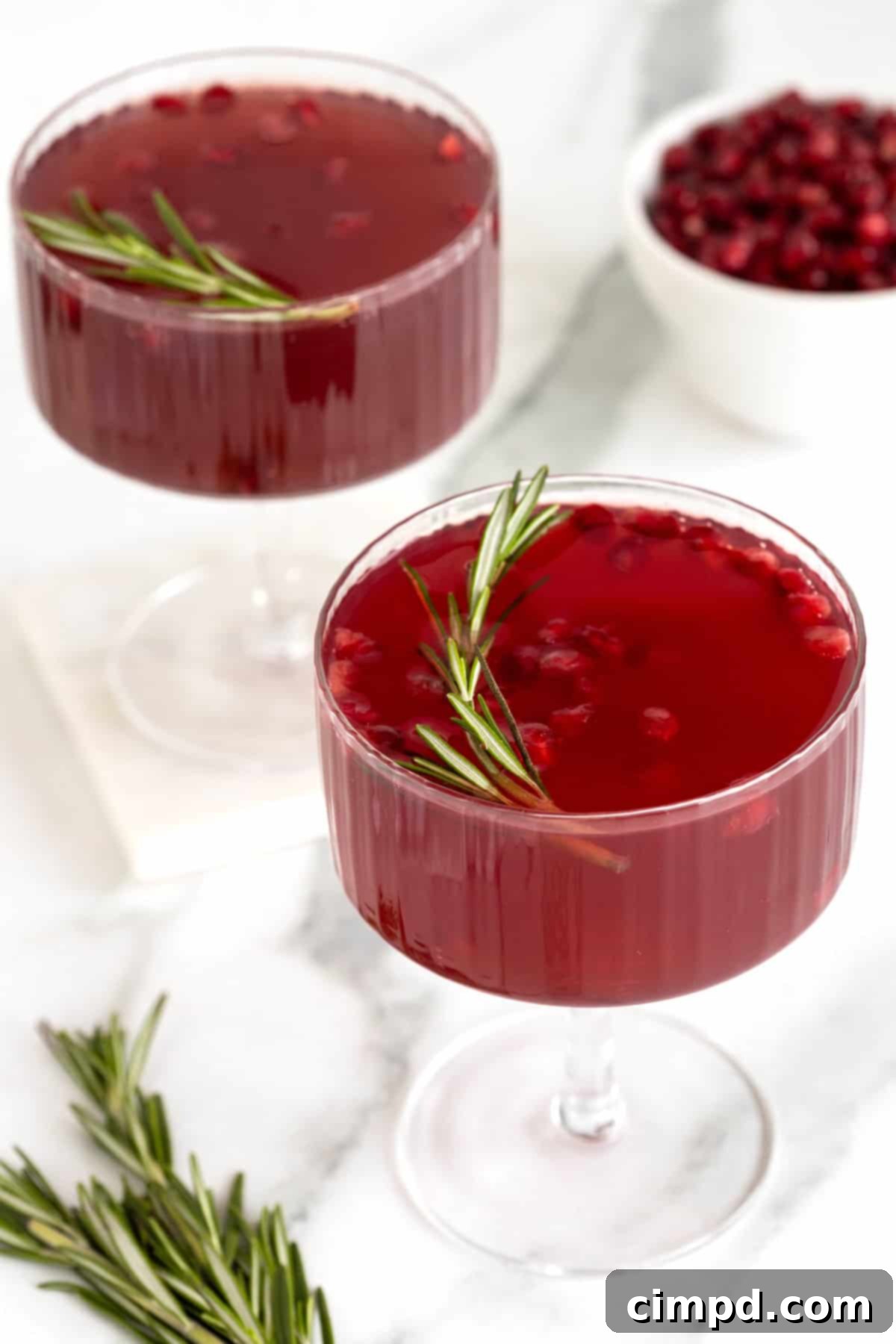 Two exquisitely prepared Pomegranate Rosemary Holiday Cocktails in elegant coupe glasses, adorned with glistening pomegranate seeds and fragrant rosemary sprigs, ready for a festive toast.