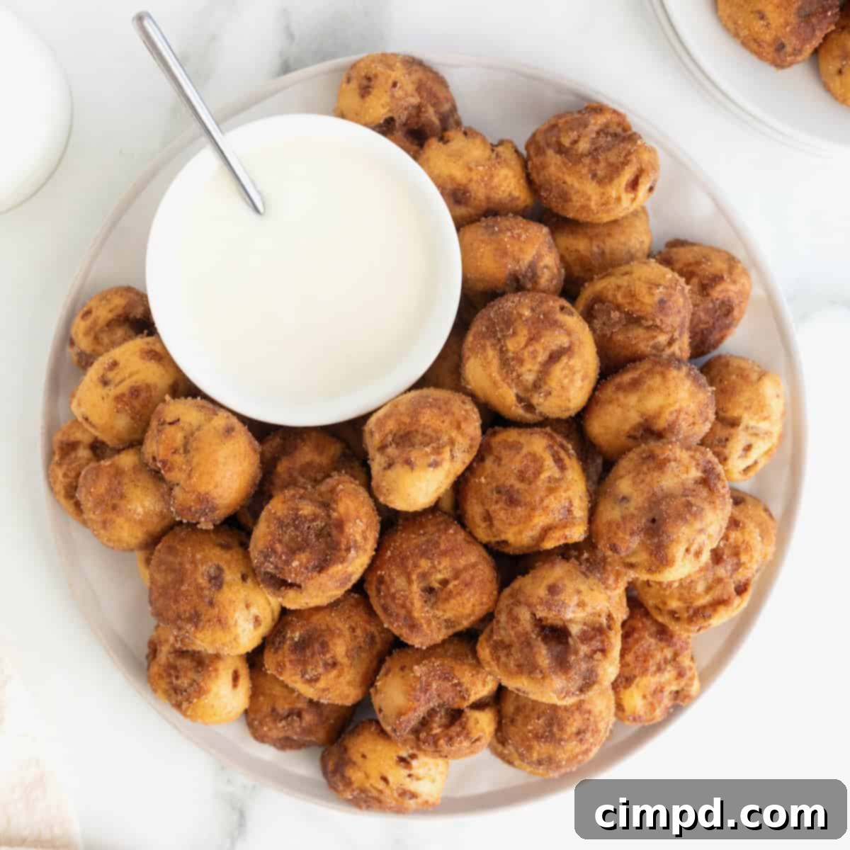 A large white serving plate piled high with golden brown cinnamon roll bites, ready for sharing. A small white bowl of creamy white icing sits invitingly next to the bites, perfect for dipping.