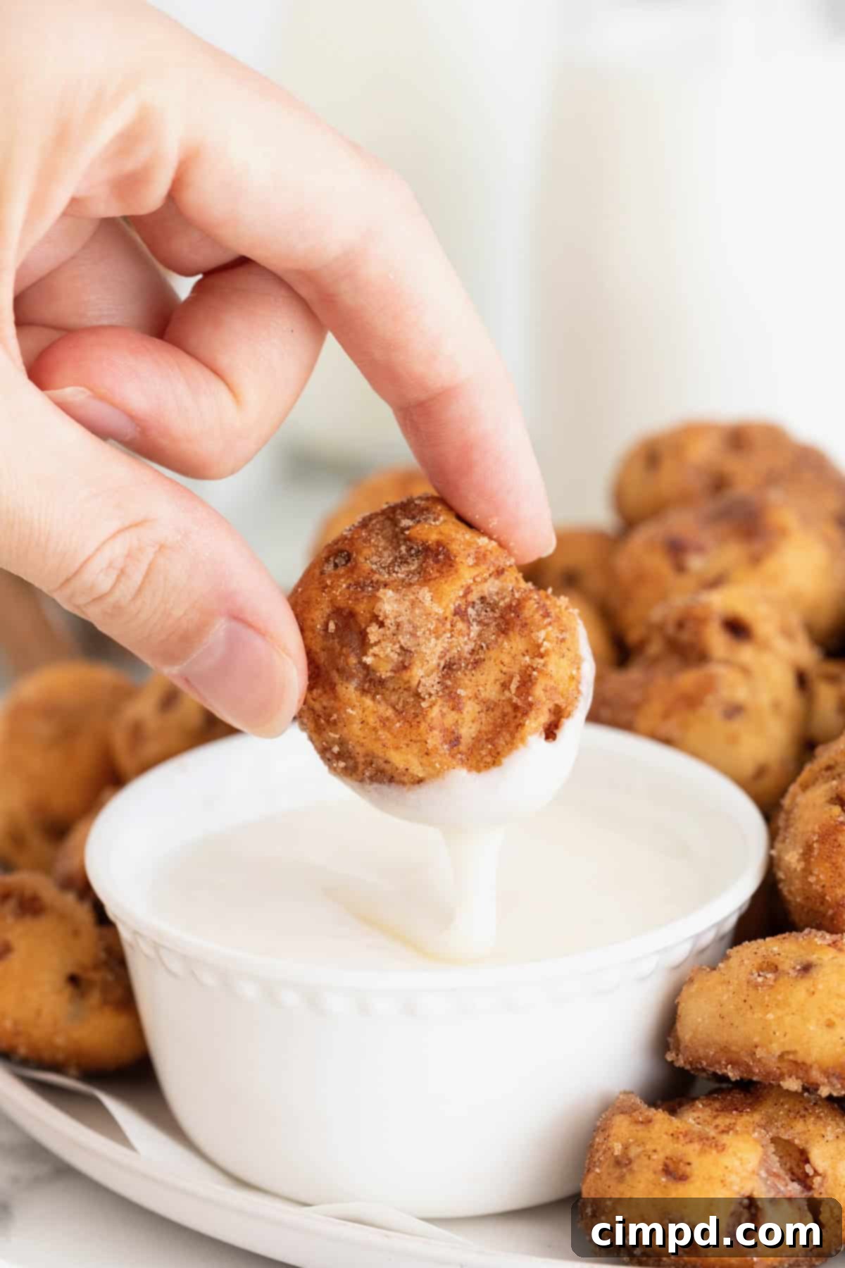 A close-up of a hand gently holding a warm cinnamon roll bite, poised to dip it into a luscious bowl of sweet white icing. The golden brown exterior of the bite is visible, dusted with cinnamon sugar.