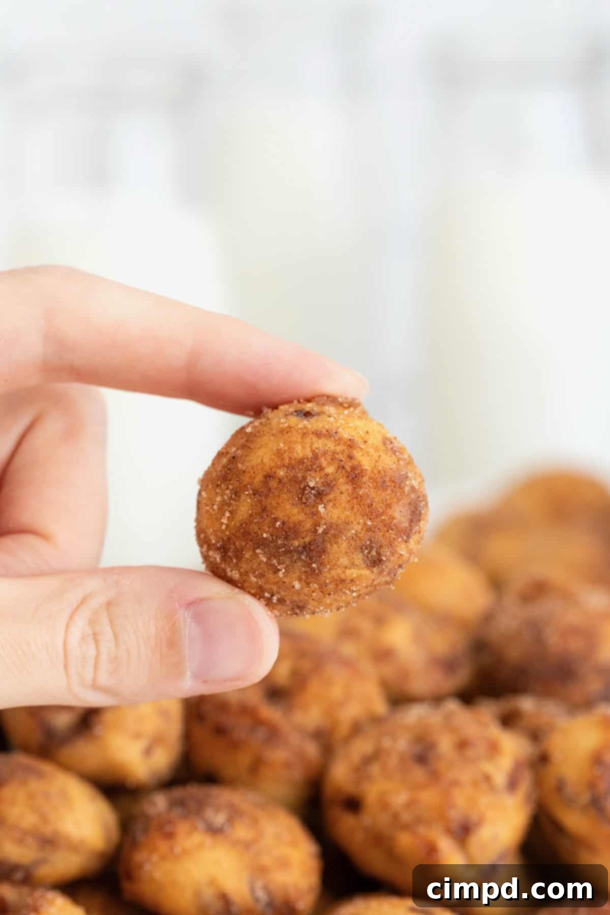 A hand holds a perfectly golden brown cinnamon roll bite between the thumb and forefinger, showcasing its sugary coating and inviting texture.