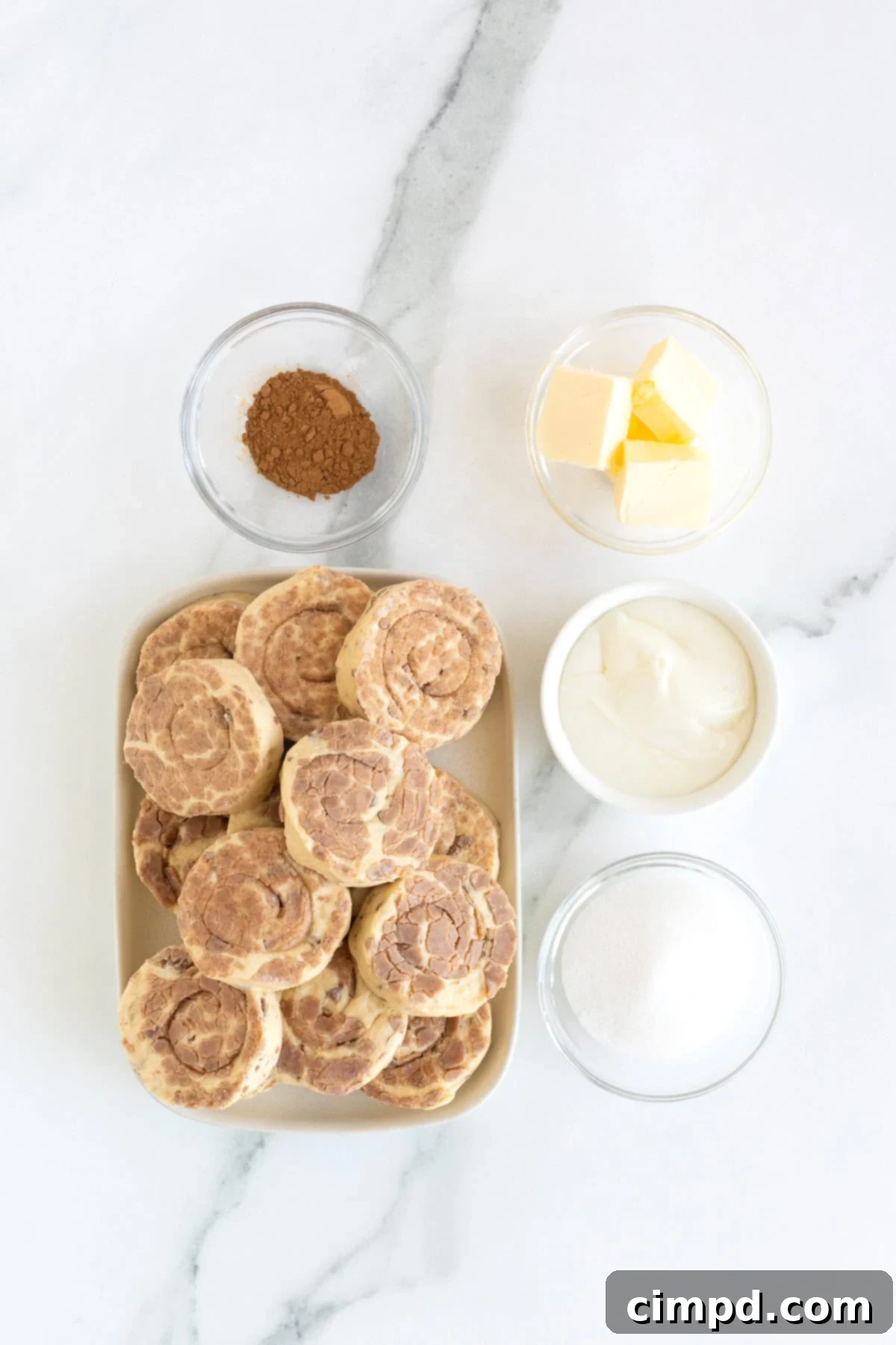 A perfectly arranged shot of the simple ingredients needed for cinnamon roll bites: canned cinnamon rolls, a stick of butter, granulated sugar, and ground cinnamon, all neatly presented in glass dishes on a white marble counter.