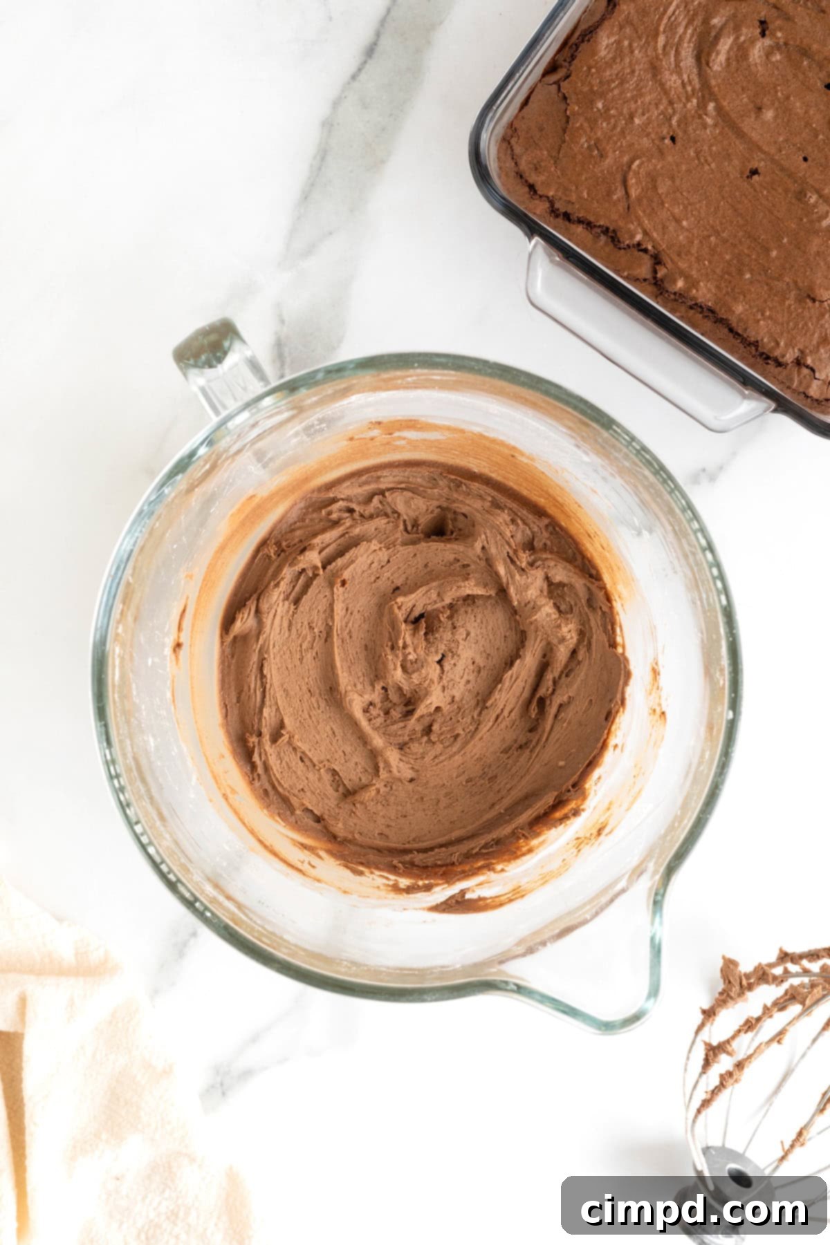 Mocha Velvet Brownies 10 A glass mixing bowl of chocolate frosting on a white marble counter.
