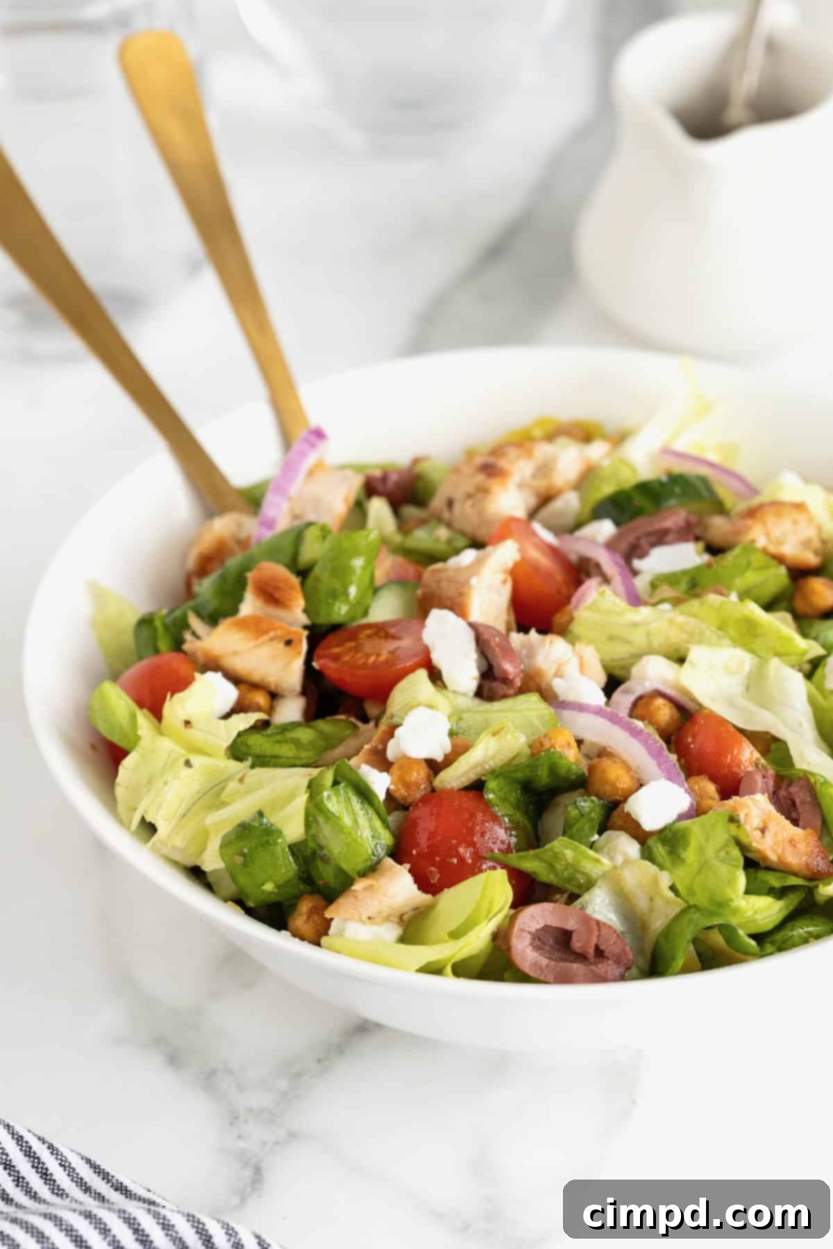 A generously sized white serving bowl filled with a colorful Greek salad, topped with perfectly grilled marinated chicken, bright red cherry tomatoes, thinly sliced Kalamata olives, crumbled feta cheese, and golden roasted chickpeas.