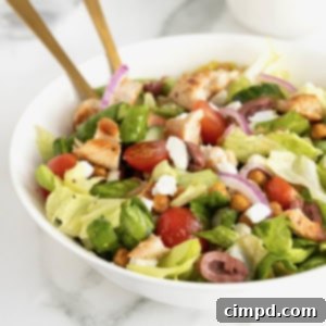 A large white serving bowl with greek salad topped with chicken, tomatoes, sliced greek olives, feta, and roasted chickpeas.