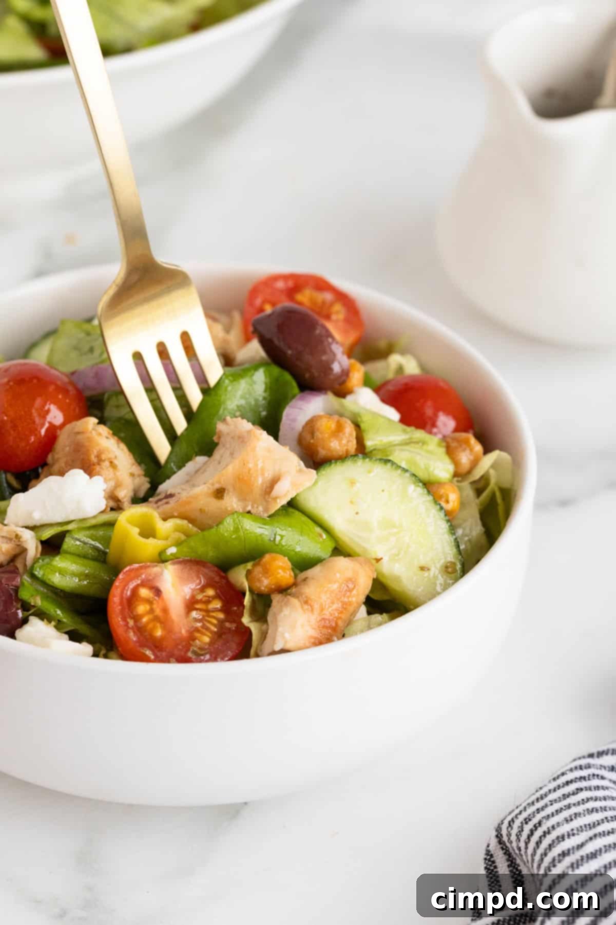 A close-up shot of a white bowl overflowing with Greek salad ingredients, including glistening cherry tomatoes, sliced Greek olives, crumbled feta, golden roasted chickpeas, and succulent pieces of marinated chicken.