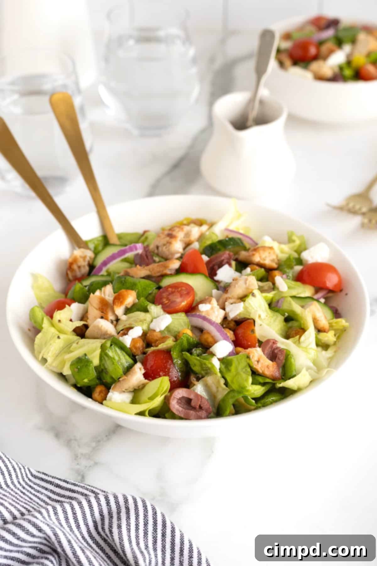 A large white serving bowl showcases a beautifully arranged Greek salad, generously topped with tender chicken, vibrant cherry tomatoes, sliced Kalamata olives, creamy feta, and crispy roasted chickpeas, ready to be served.