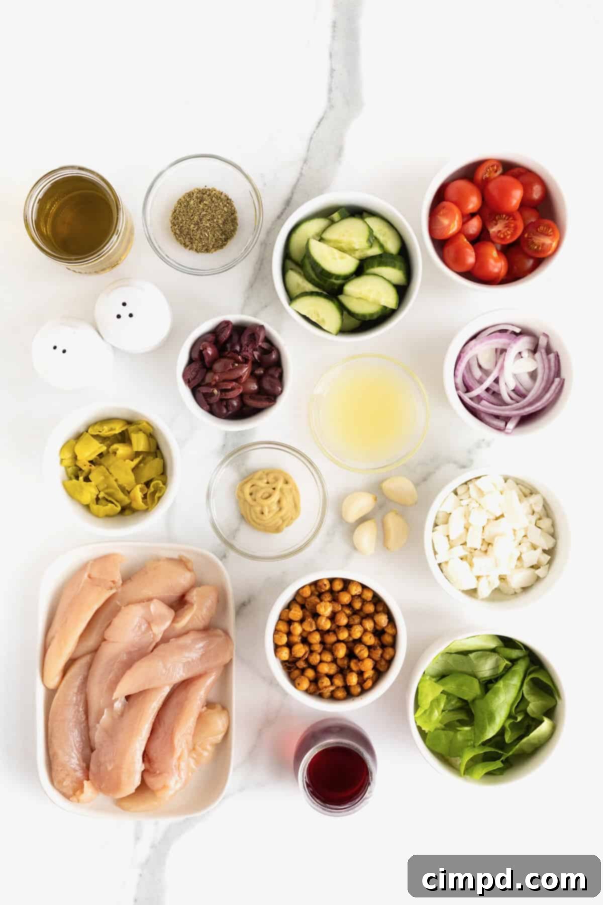 Various ingredients for Greek Salad with Marinated Chicken, including fresh vegetables, herbs, and oils, artfully arranged in small glass dishes on a pristine white marble counter, ready for preparation.