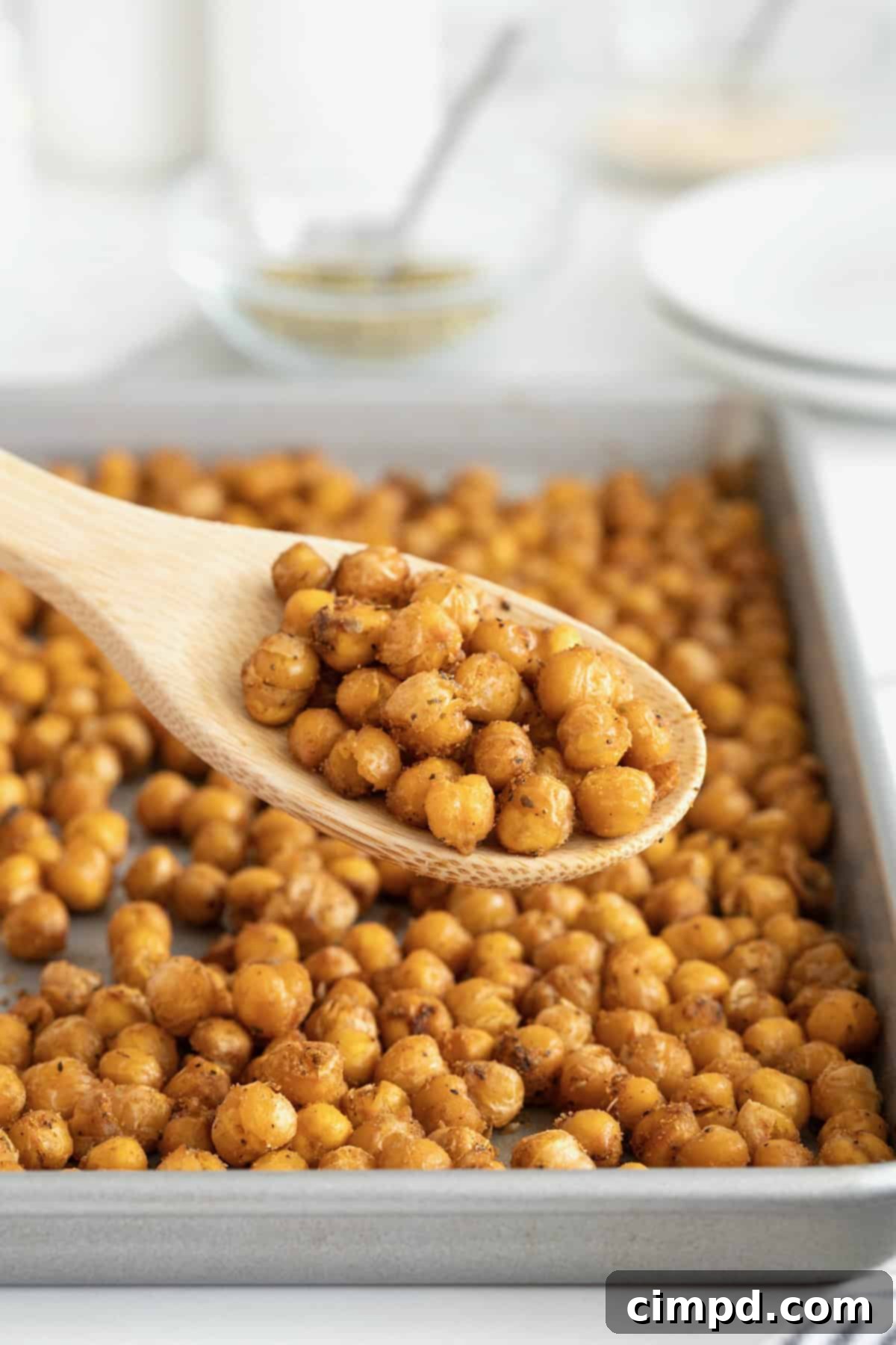 A rustic wooden spoon brimming with uncooked chickpeas, poised in front of a gleaming aluminum baking sheet generously spread with perfectly roasted, golden-brown chickpeas. This image captures the journey from raw ingredient to delicious, crispy snack.
