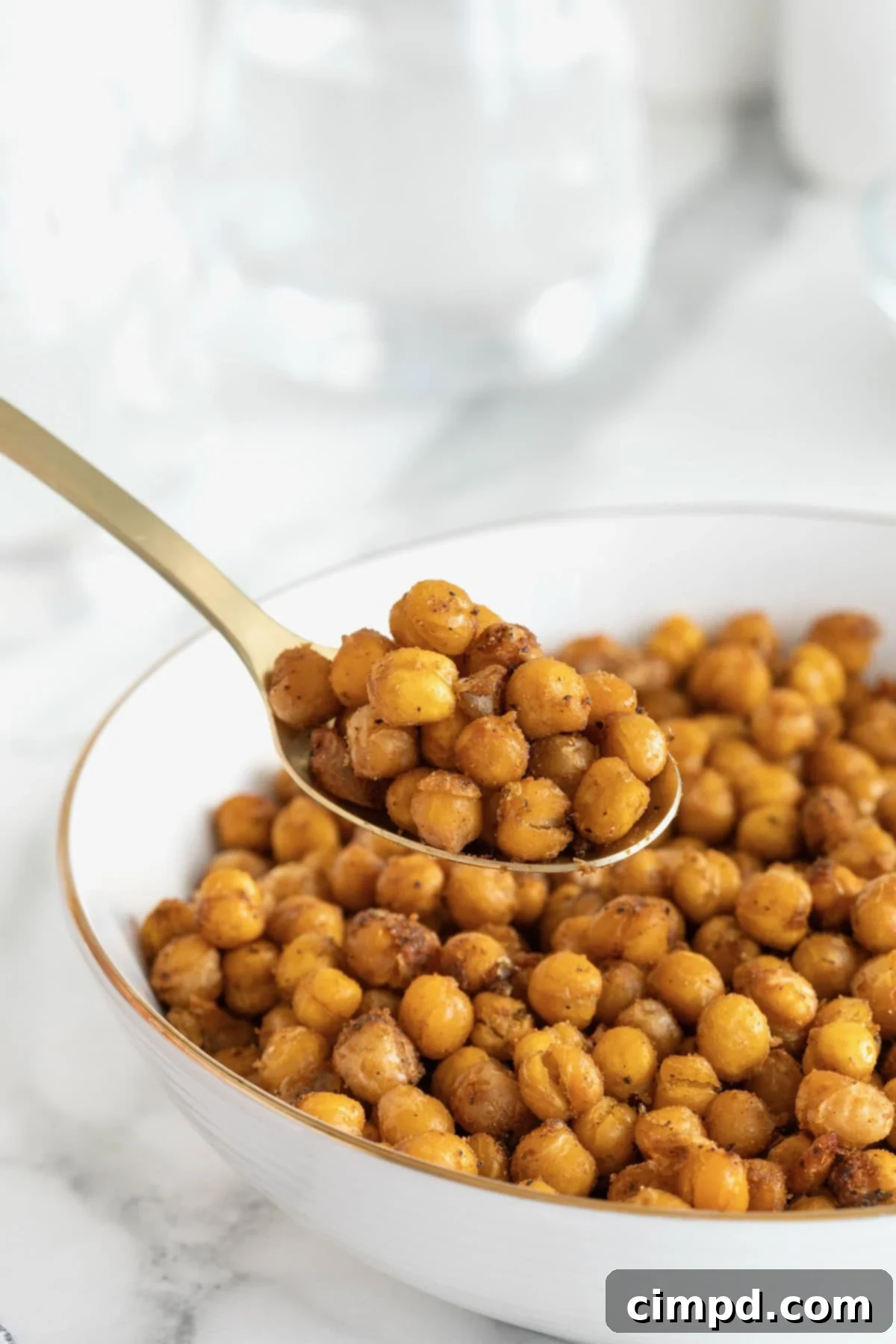 A gleaming silver spoon elegantly presents a heap of golden-brown roasted chickpeas, artfully suspended above a stylish white serving bowl. The chickpeas glisten with savory seasoning, highlighting their irresistible crispness.