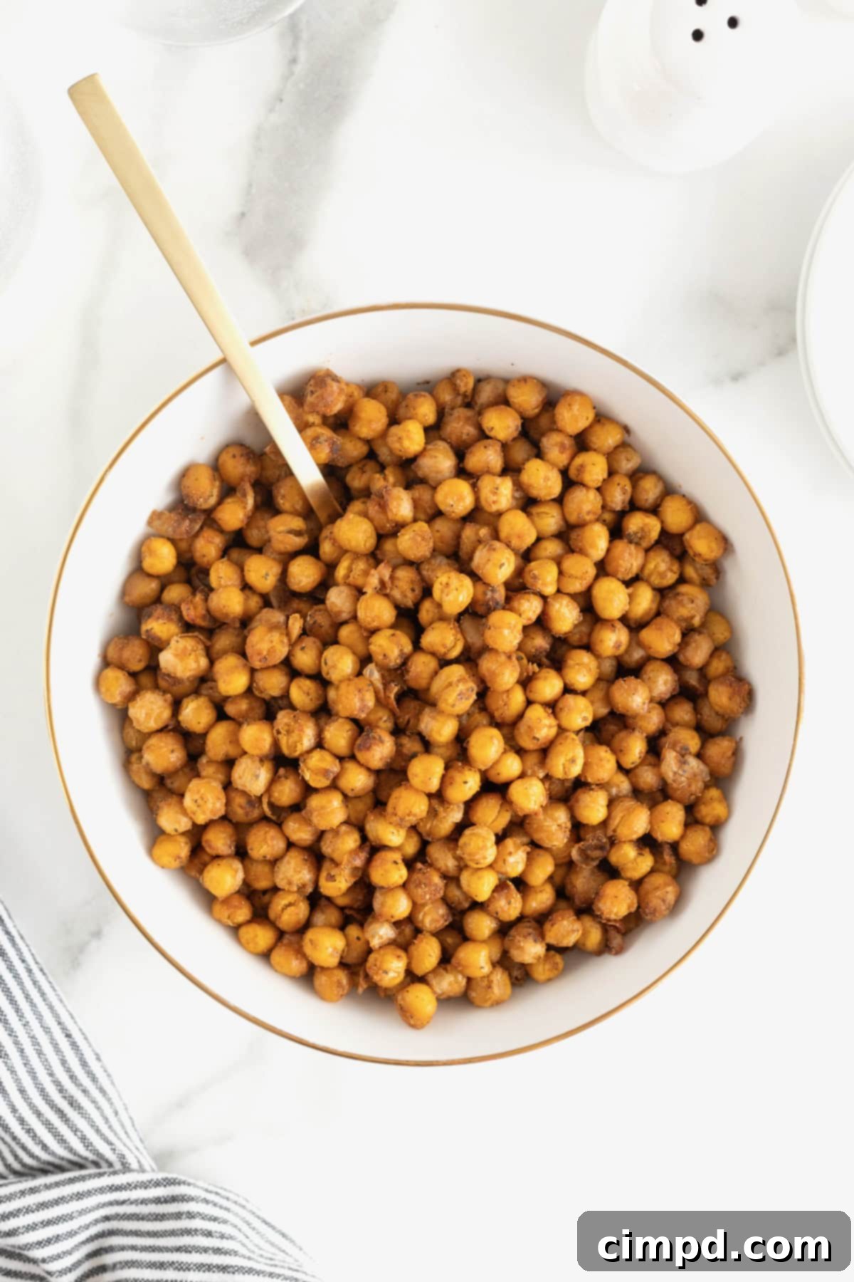 A pristine, large white serving bowl is filled to the brim with perfectly roasted, golden-brown chickpeas, generously seasoned. The bowl rests invitingly on a cool, white marble kitchen counter, suggesting a wholesome and delicious snack or meal component.