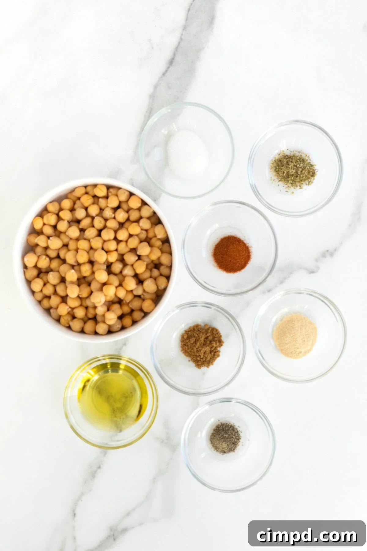 A collection of small, elegant glass dishes meticulously arranged on a pristine white marble counter. Each dish holds a distinct ingredient for crafting roasted chickpeas: extra-virgin olive oil, dried oregano, garlic powder, paprika, ground cumin, salt, and ground pepper, all awaiting preparation.