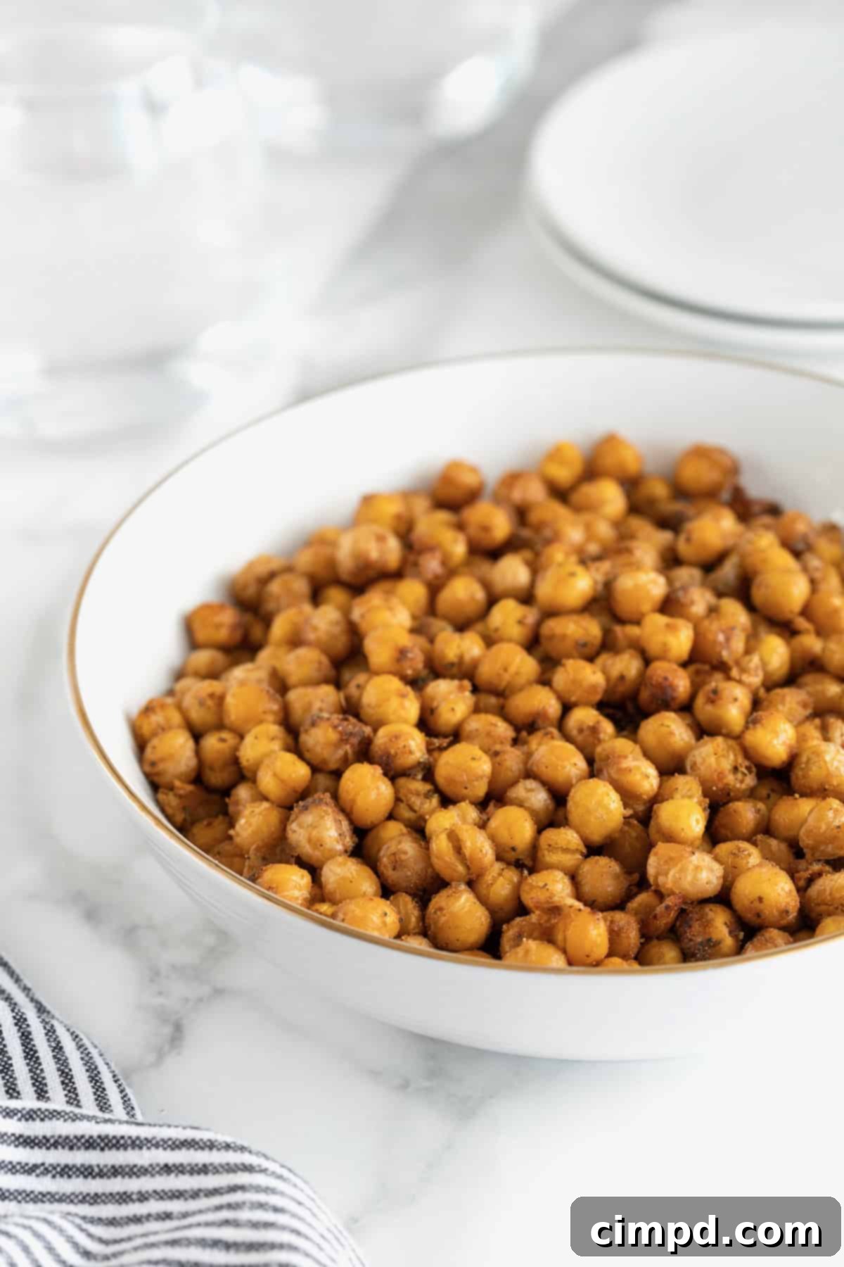A pristine, large white serving bowl is filled to the brim with perfectly roasted, golden-brown chickpeas, generously seasoned. The bowl rests invitingly on a cool, white marble kitchen counter, ready to be served or enjoyed as a healthy snack.