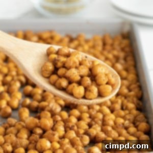 A wooden spoon of chickpeas in front of an aluminum baking sheet of roasted chickpeas, showing the crisp texture.