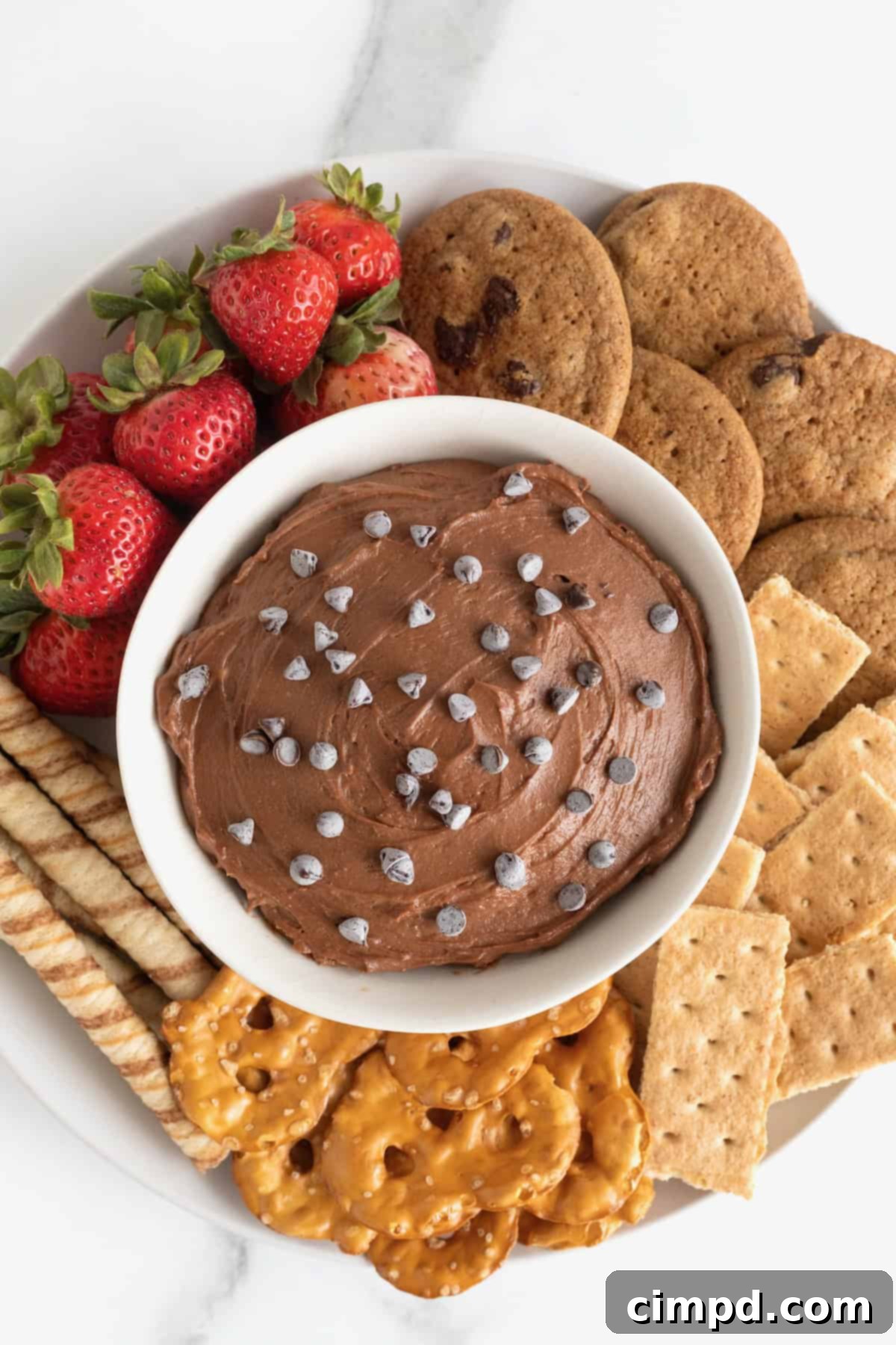 Decadent Chocolate Cheesecake Dip 8 A beautifully presented white bowl of chocolate cheesecake dip, generously topped with mini chocolate chips and artfully surrounded by a variety of dippers, including fresh strawberries, different types of cookies, and savory pretzels.