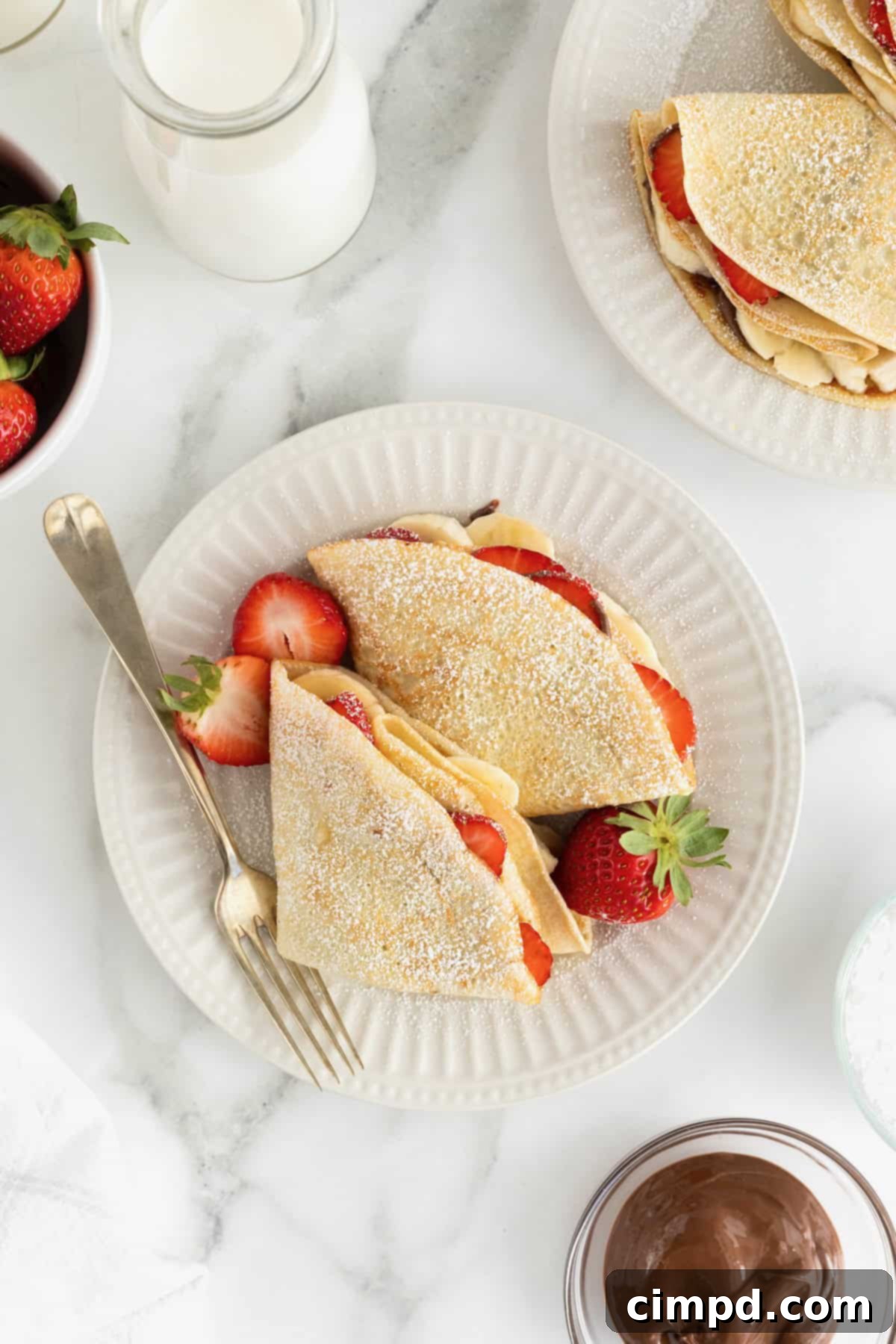Two perfectly folded sweet crepes, filled with fresh strawberry and banana slices, drizzled with Nutella, and lightly dusted with powdered sugar, resting on an elegant white fluted plate with a fork beside them. These delicious crepes are ready to be enjoyed.