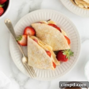 Two crepes on a white fluted plate with a fork. The crepes are filled with strawberry slices and banana slices and dusted with powdered sugar.