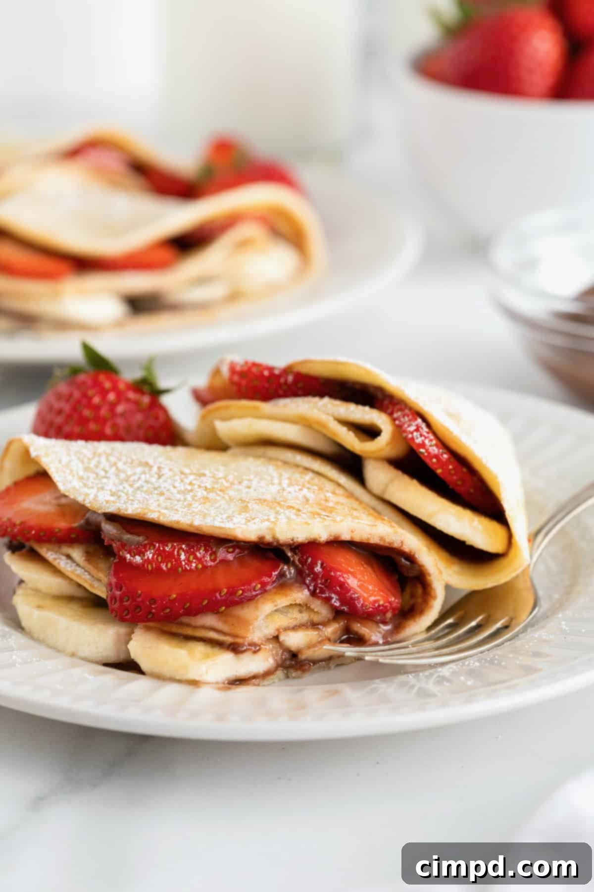 Close-up of two elegantly folded sweet crepes, overflowing with fresh strawberry and banana slices, drizzled with rich Nutella, and lightly dusted with powdered sugar. They are presented on a pristine white fluted plate, ready for serving.