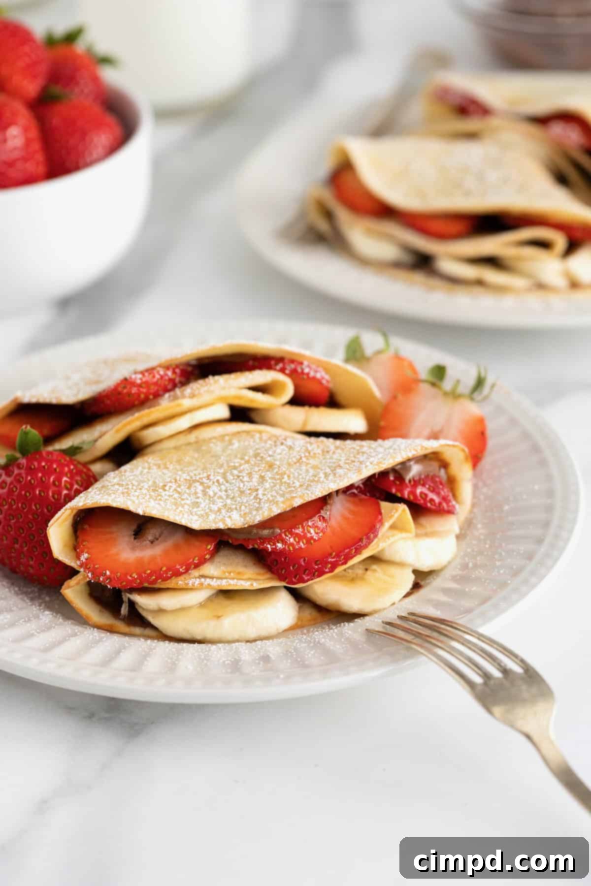 A close-up view of two delicate sweet crepes, artistically folded into quarters and filled with a harmonious blend of strawberry and banana slices, and rich Nutella. They are elegantly presented on a white fluted plate, awaiting a final dusting of powdered sugar.