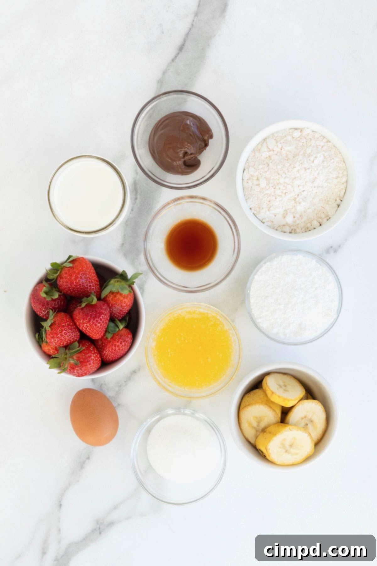 A collection of fresh ingredients for sweet crepes, including flour, milk, eggs, sugar, melted butter, and vanilla extract, neatly arranged in small glass bowls on a white marble countertop.
