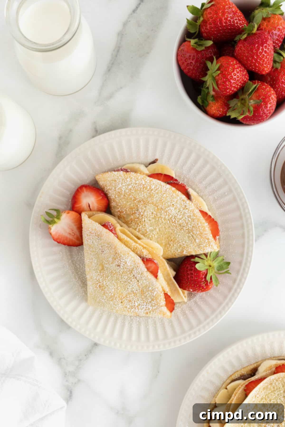 Two freshly prepared sweet crepes, elegantly folded and generously dusted with powdered sugar, presented on a white fluted plate.