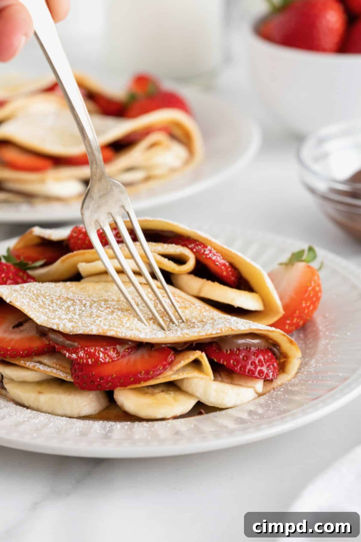 A beautiful presentation of two sweet crepes, filled with Nutella, strawberries, and bananas, and generously dusted with powdered sugar, arranged on a pristine white fluted plate. The image highlights the inviting texture and vibrant colors of the fresh fruit.