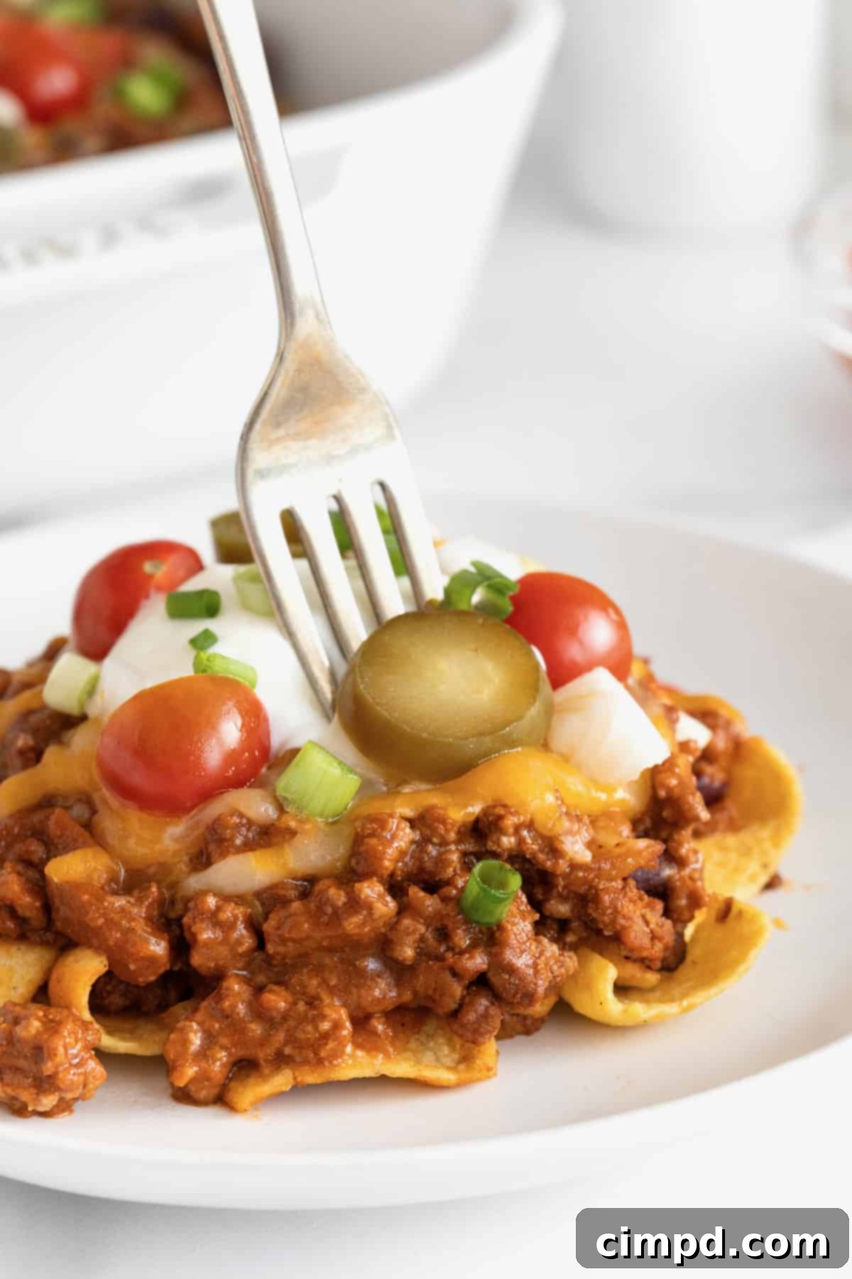 Loaded Frito Chili Bake 3 A generous serving of Frito chili pie casserole on a white plate with a metal fork sticking out, showcasing its layers.
