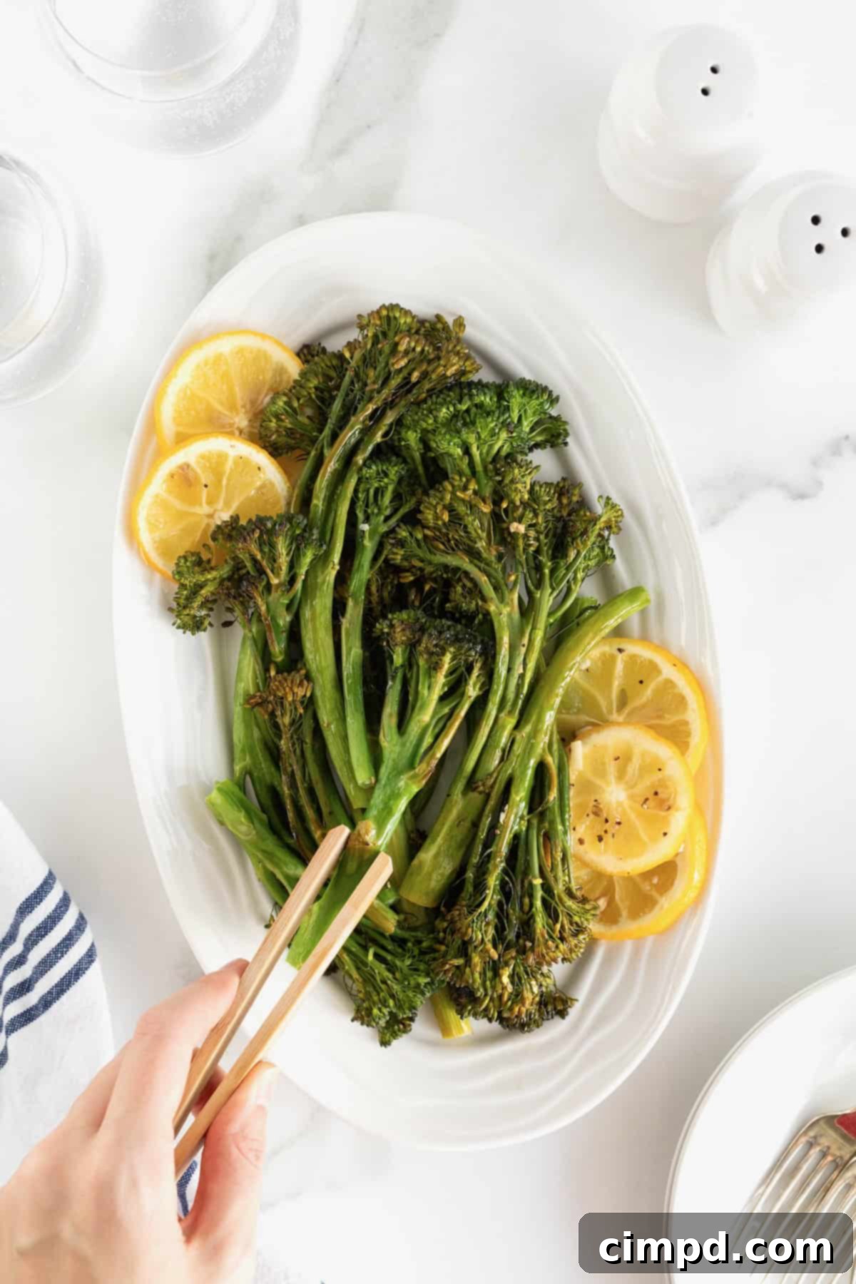 A piece of roasted broccolini being held by wooden tongs over a plate of broccolini, showcasing its perfect char and tenderness.