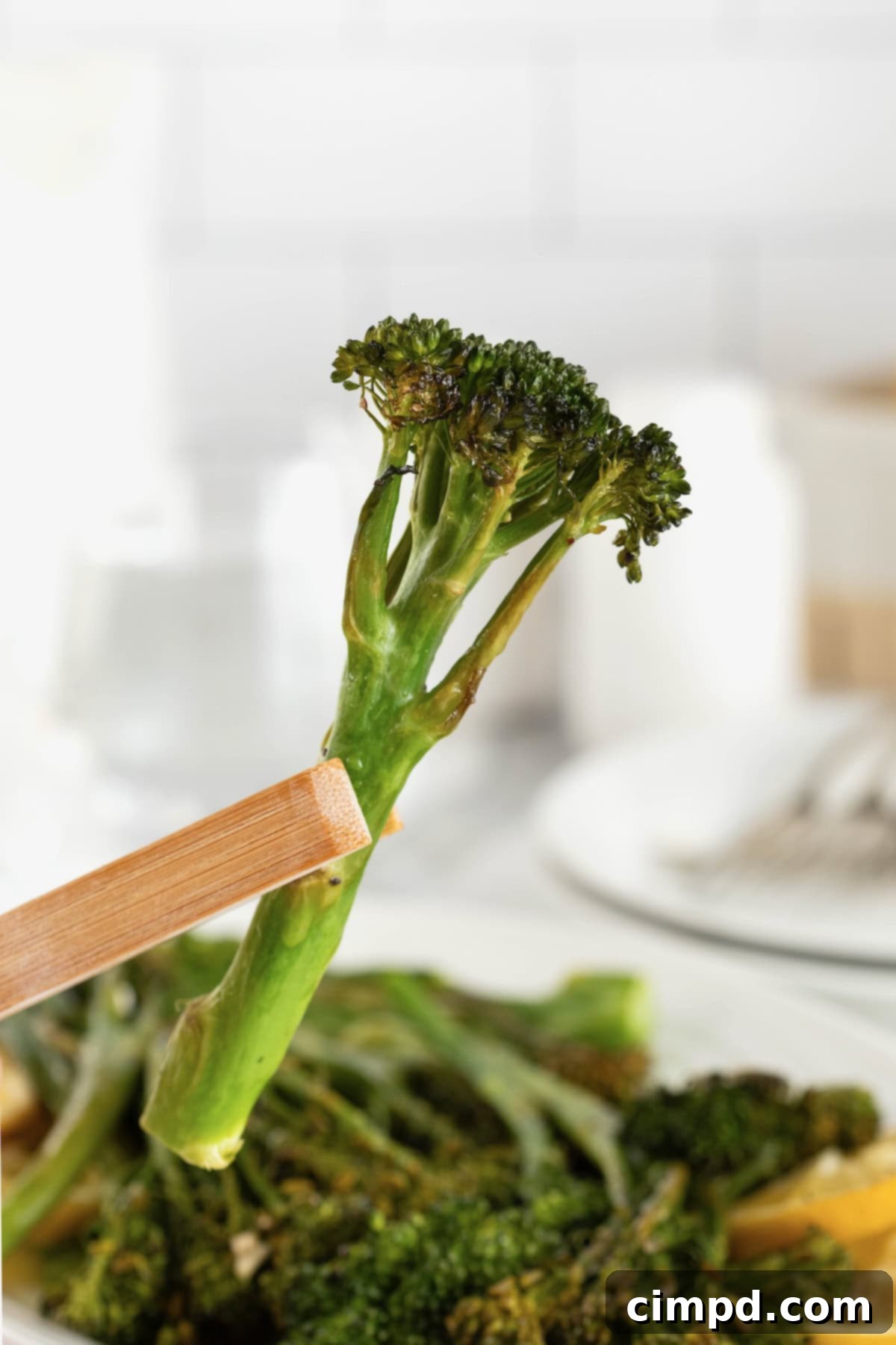 A close-up shot of a piece of roasted broccolini being held by wooden tongs over a plate of broccolini, highlighting its vibrant green color and perfect char.