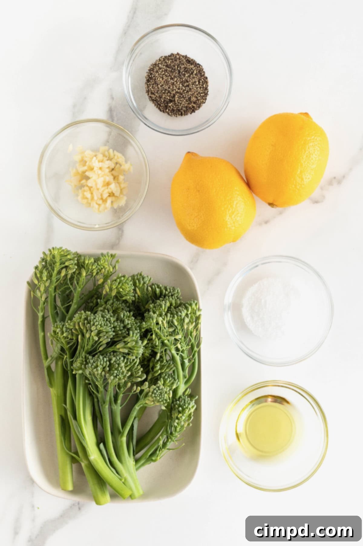 Various ingredients for roasted broccolini, including fresh broccolini, lemons, garlic cloves, olive oil, salt, and pepper, arranged in small glass dishes on a white marble counter.