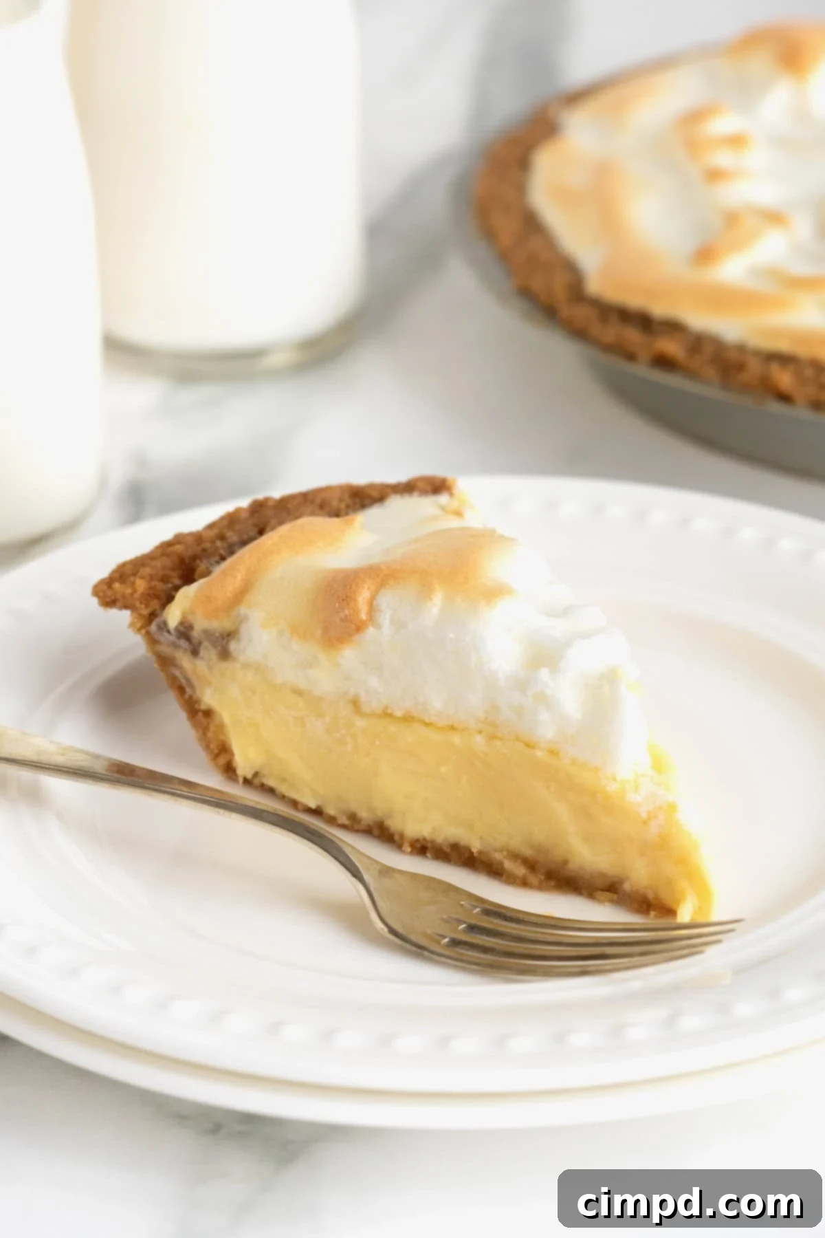 Zesty Meringue Dream 2 A beautifully plated slice of Lemon Meringue Pie, showcasing its vibrant yellow custard and golden-brown meringue peaks, resting beside a fork on a white dessert plate with delicate beaded details.