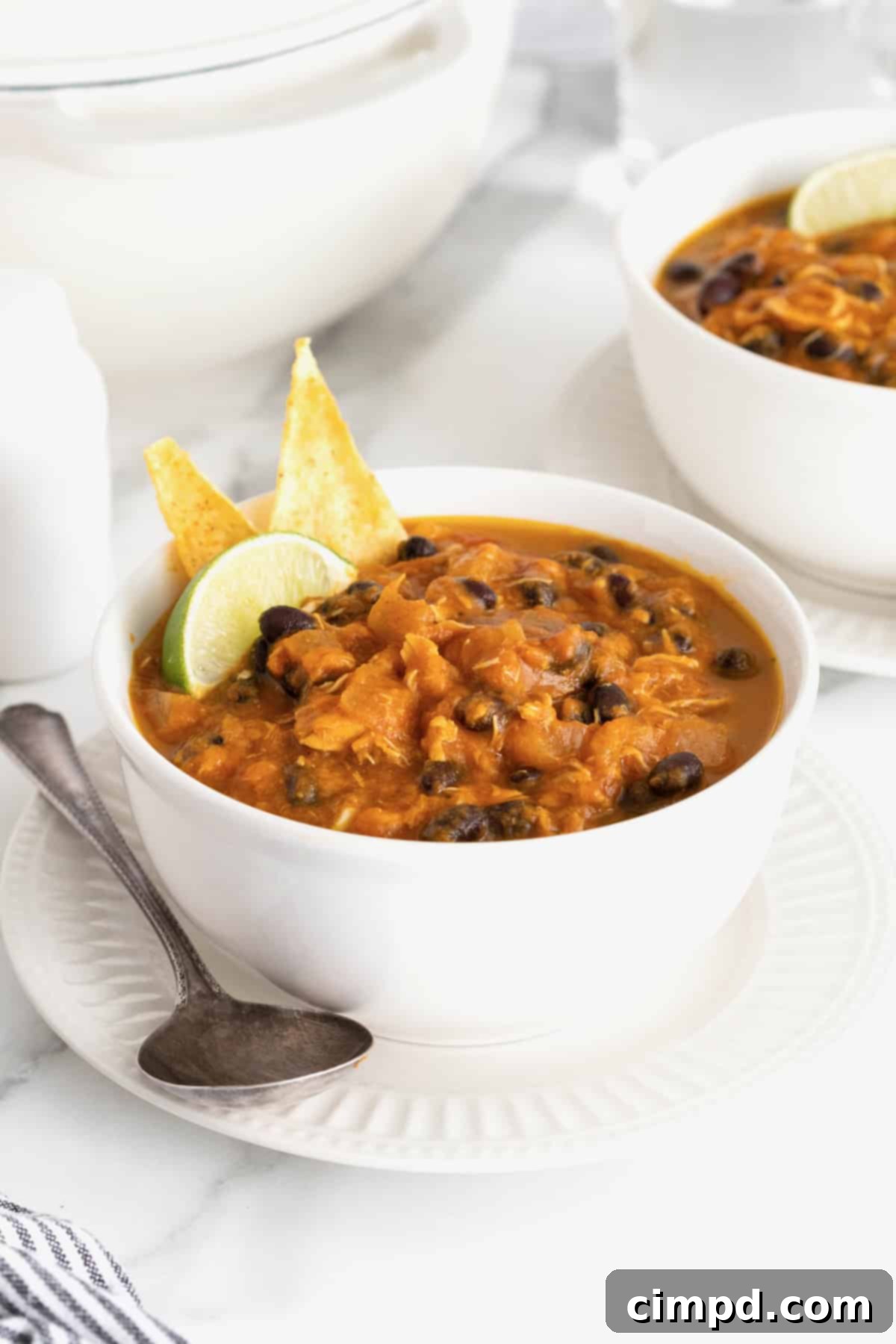 Harvest Pumpkin Chicken Chili 2 A white soup bowl of pumpkin chicken chili on a white fluted plate. There are two tortilla chips and a lime wedge garnishing the chili.