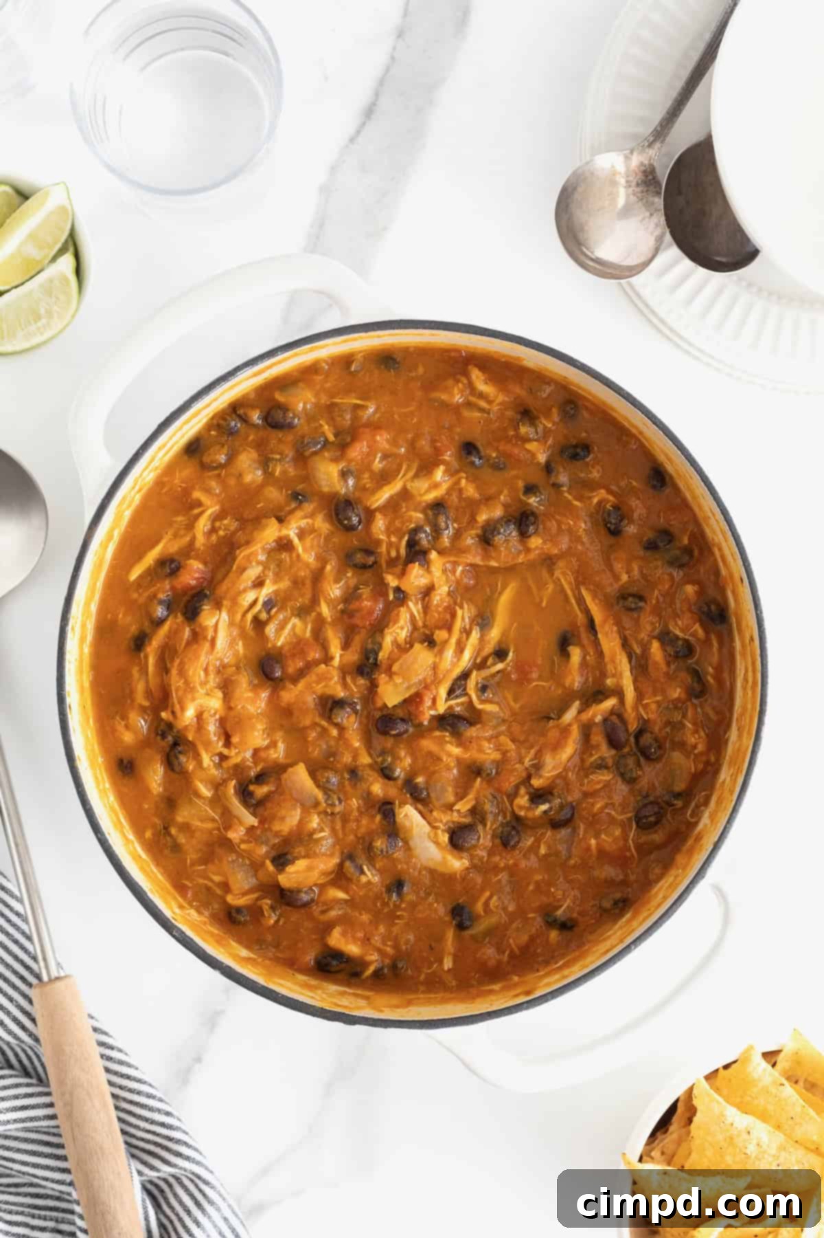 Harvest Pumpkin Chicken Chili 10 A white Dutch oven of chicken pumpkin chili on a white marble counter. A white dish of yellow corn tortilla chips sits at the bottom right.