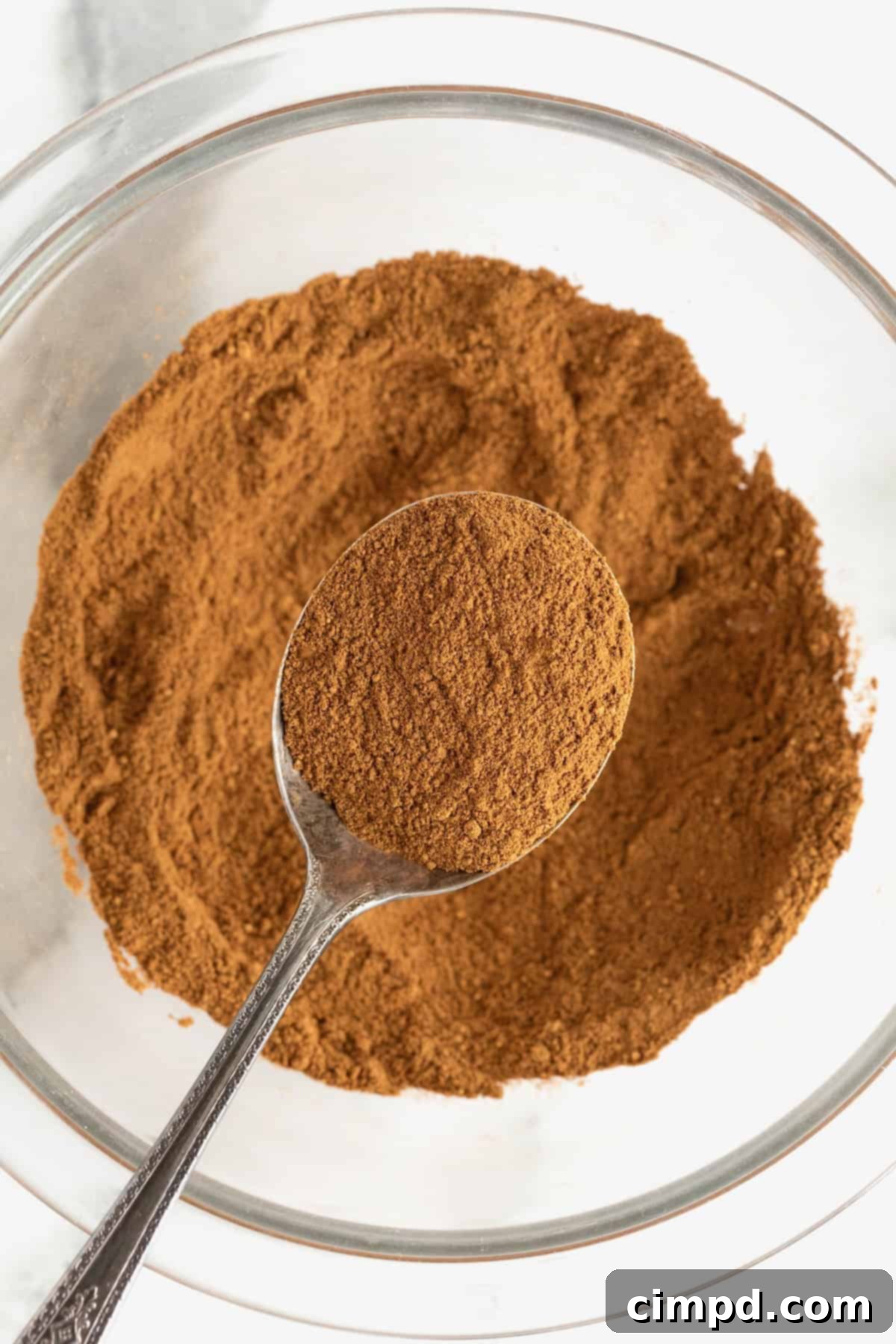 Make Your Own Pumpkin Pie Spice 2 A spoonful of vibrant, homemade pumpkin pie spice delicately balanced over a clear glass bowl, showcasing the rich blend of spices within.