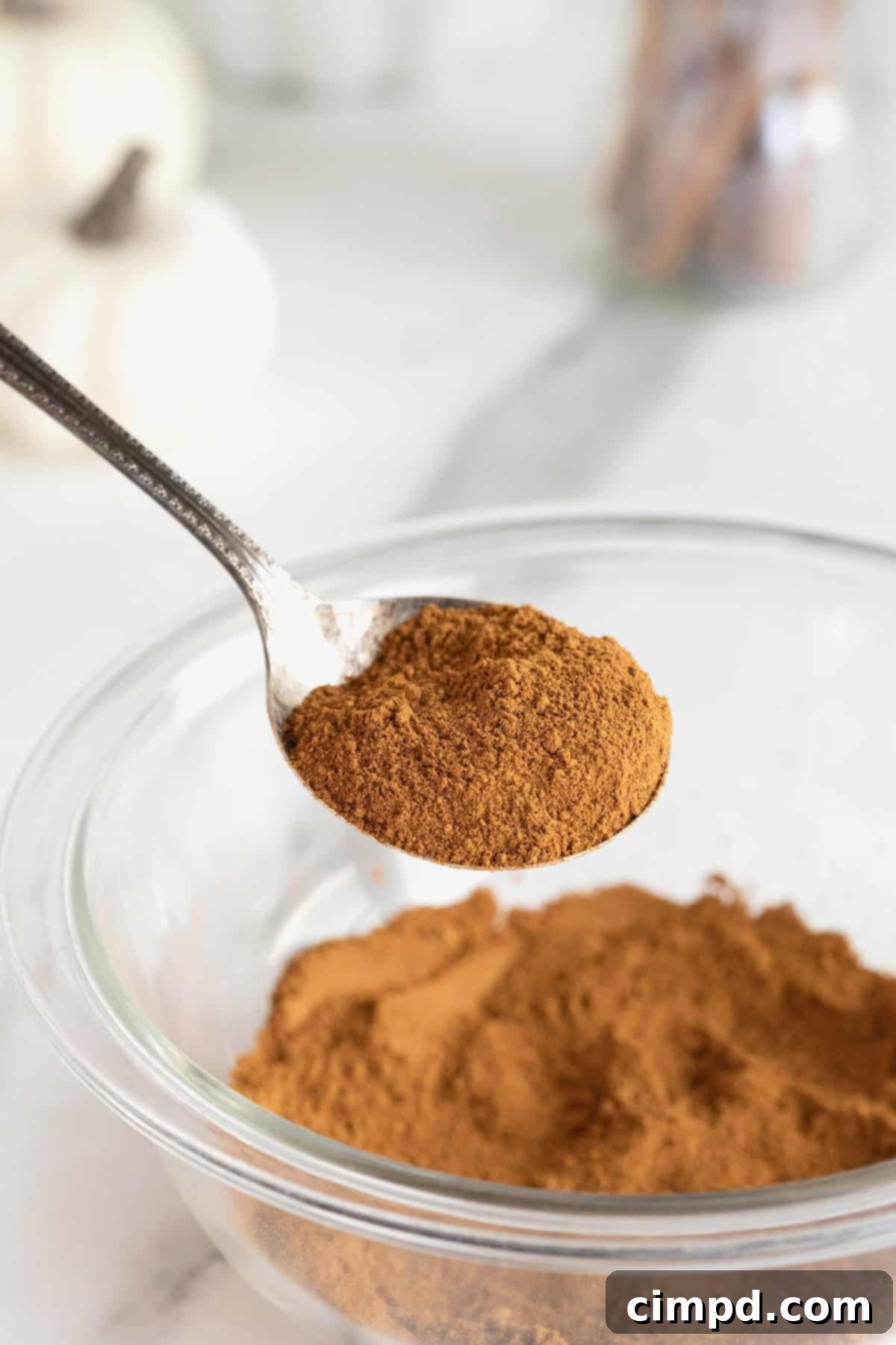 Make Your Own Pumpkin Pie Spice 4 A close-up shot of a spoon filled with freshly blended homemade pumpkin pie spice, held above a clear glass bowl, ready for baking.