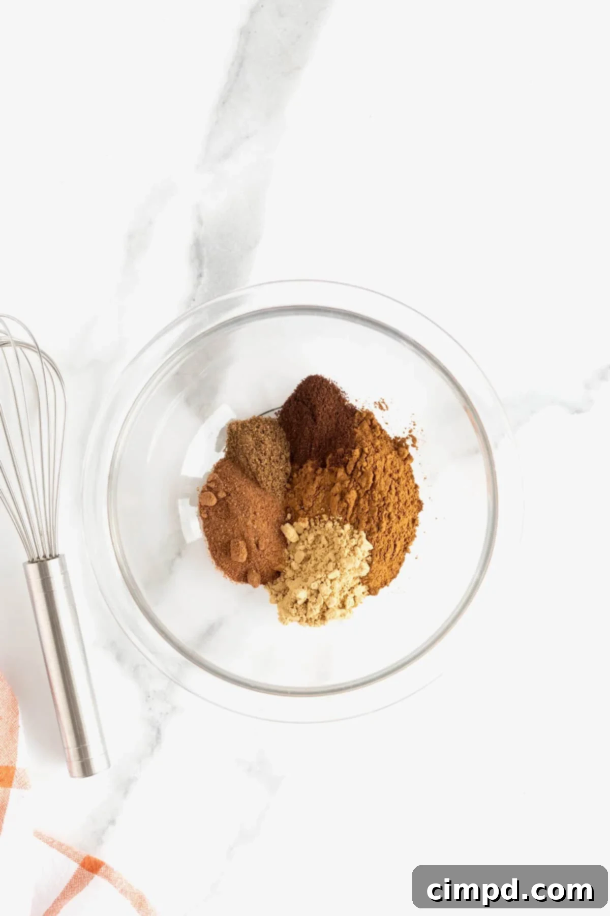 Make Your Own Pumpkin Pie Spice 5 Various ground spices, including cinnamon, nutmeg, ginger, cloves, and allspice, carefully measured and placed in a clear glass bowl on a pristine white marble counter, ready for mixing.