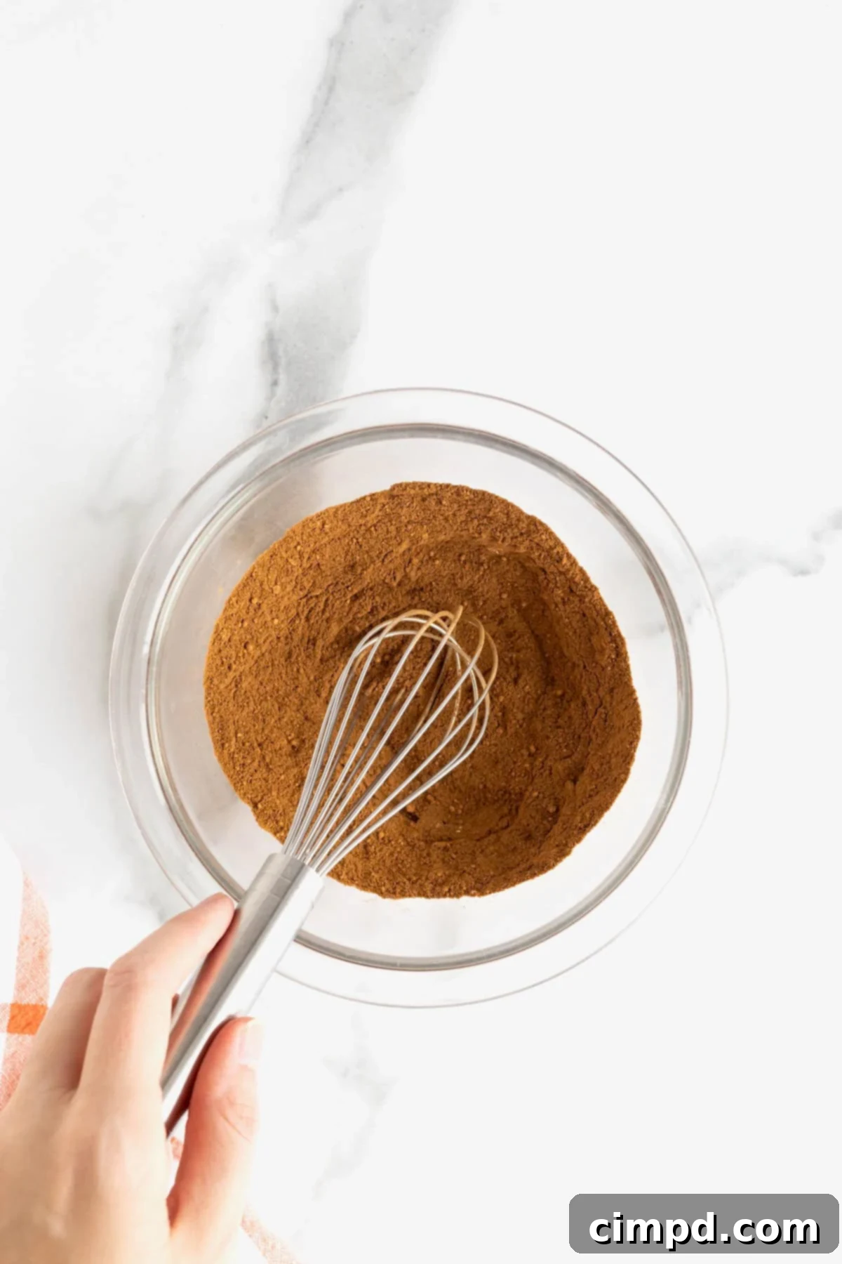 Make Your Own Pumpkin Pie Spice 6 A hand actively whisks a vibrant mixture of ground spices in a clear glass bowl, creating a swirling motion that blends them perfectly on a white marble counter.
