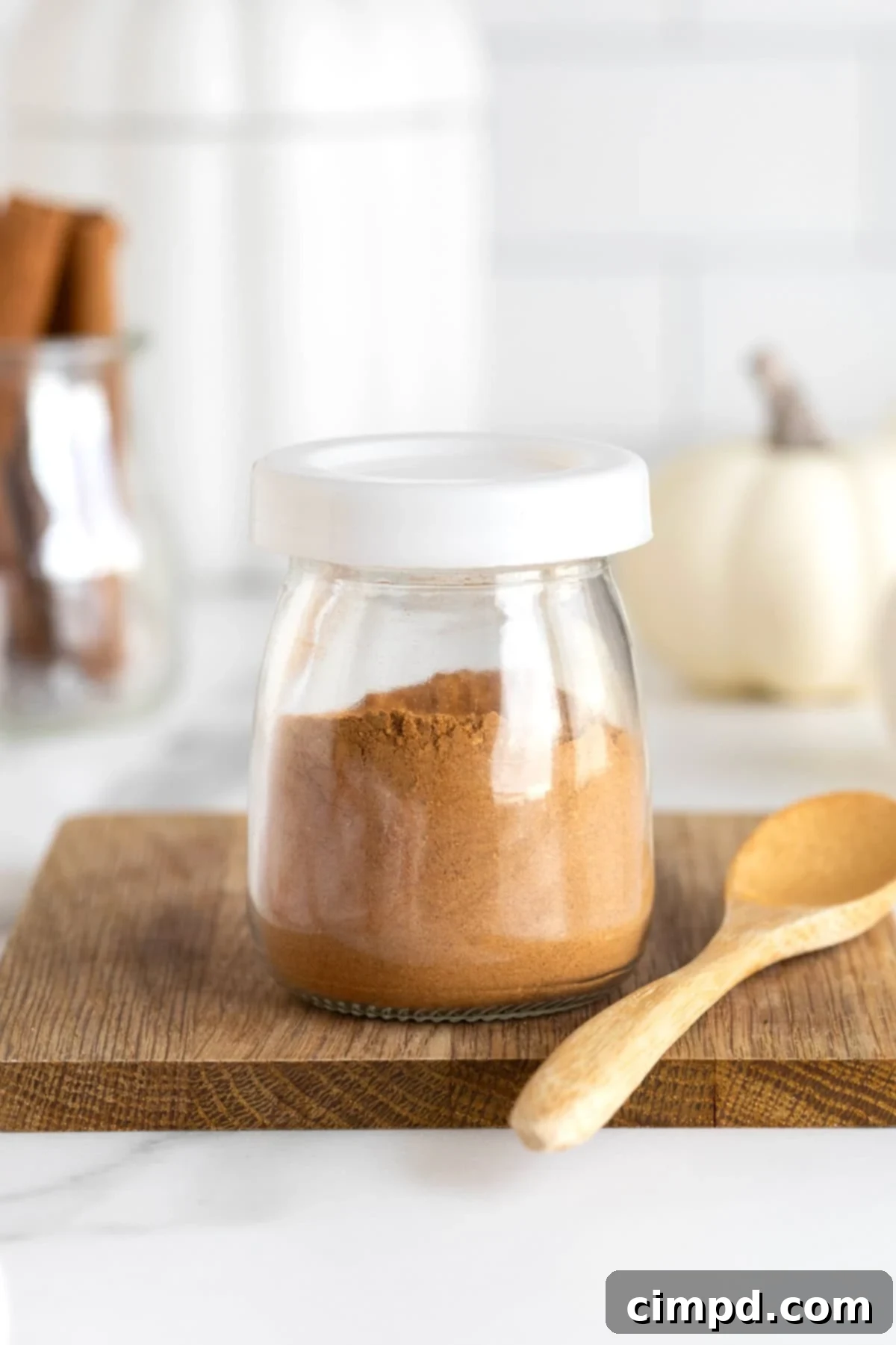 Make Your Own Pumpkin Pie Spice 7 A charming glass jar, filled to the brim with freshly prepared homemade pumpkin pie spice, sits on a rustic wooden cutting board, with a small wooden spoon nearby.