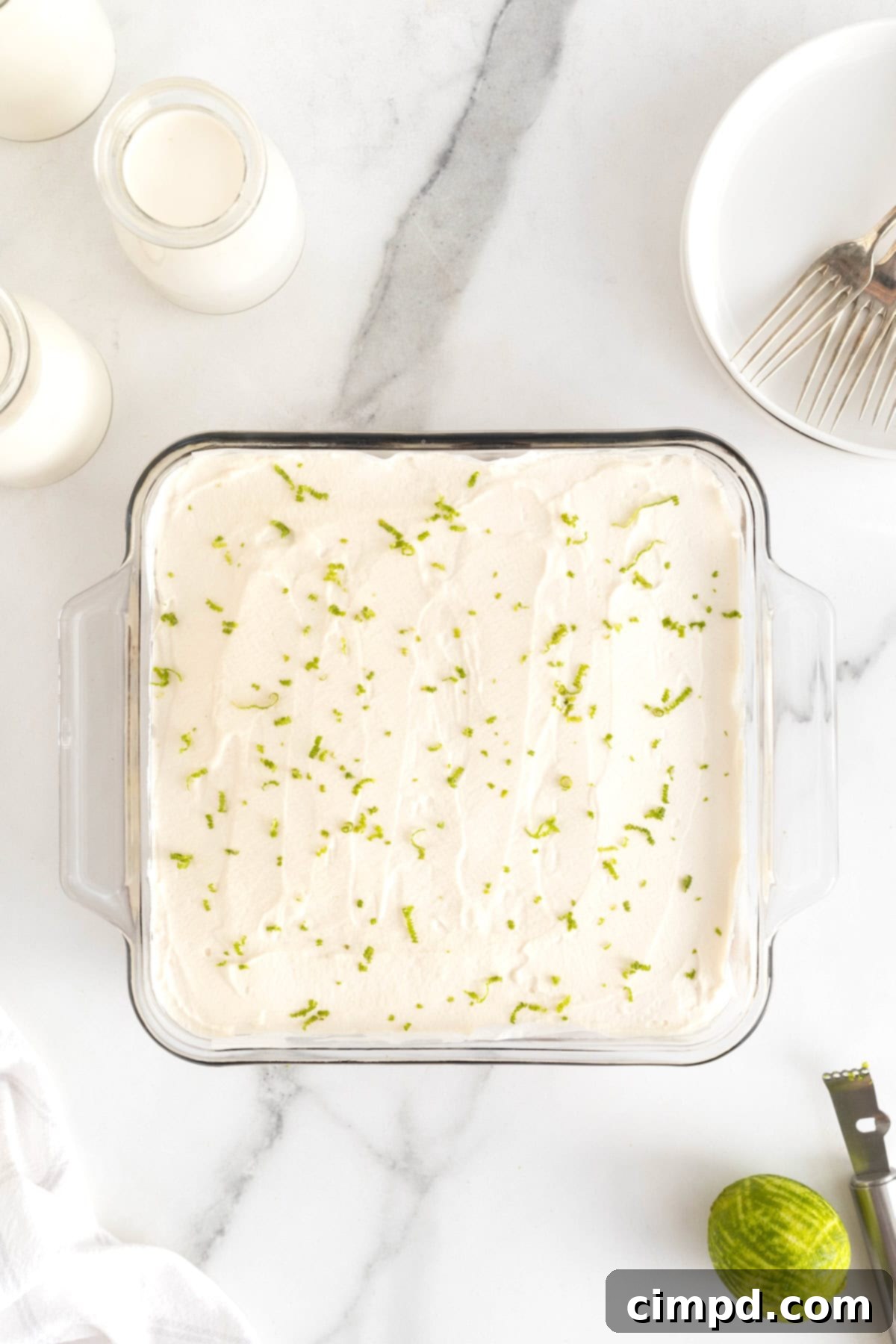 A square glass baking dish of key lime pie bars sprinkled with lime zest on a white marble counter.