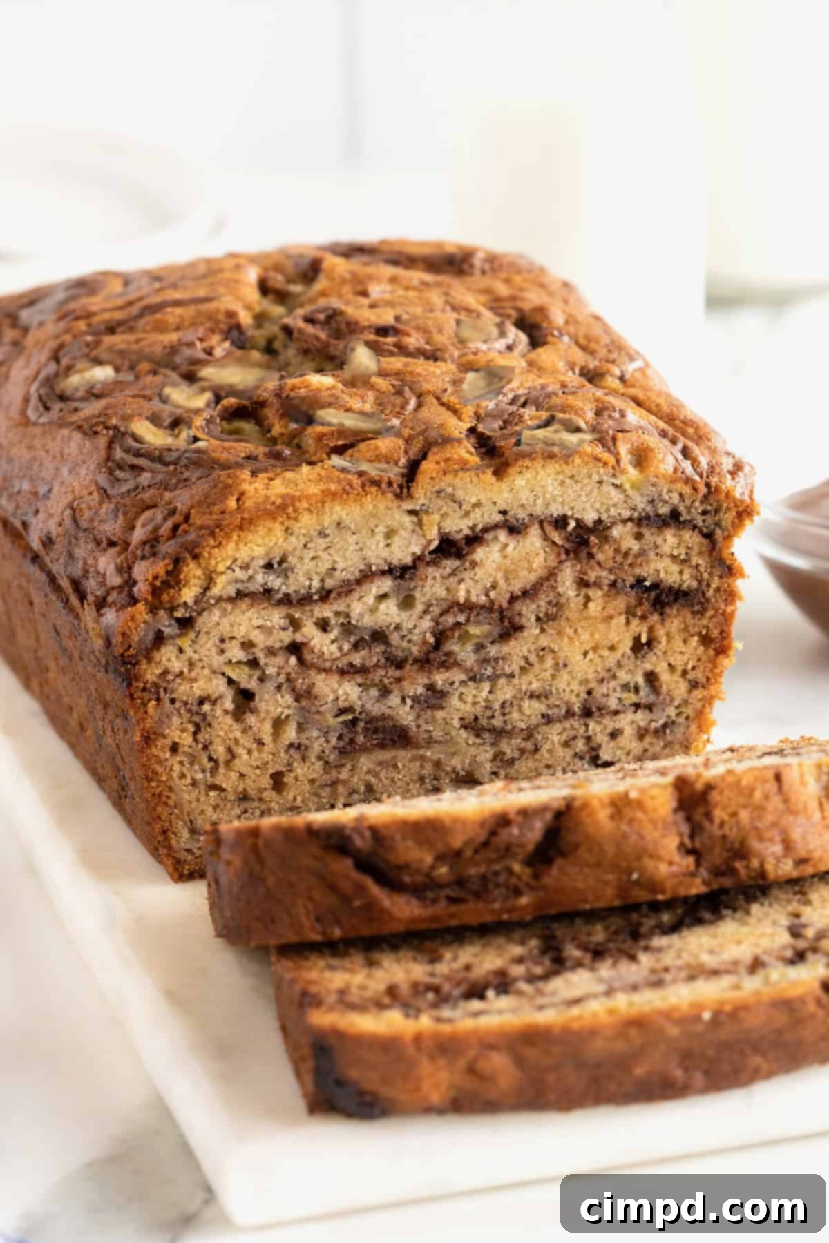 A beautifully baked loaf of Nutella swirled banana bread resting on a rustic wooden board, with several slices cut, revealing the distinctive chocolate-hazelnut swirls inside.
