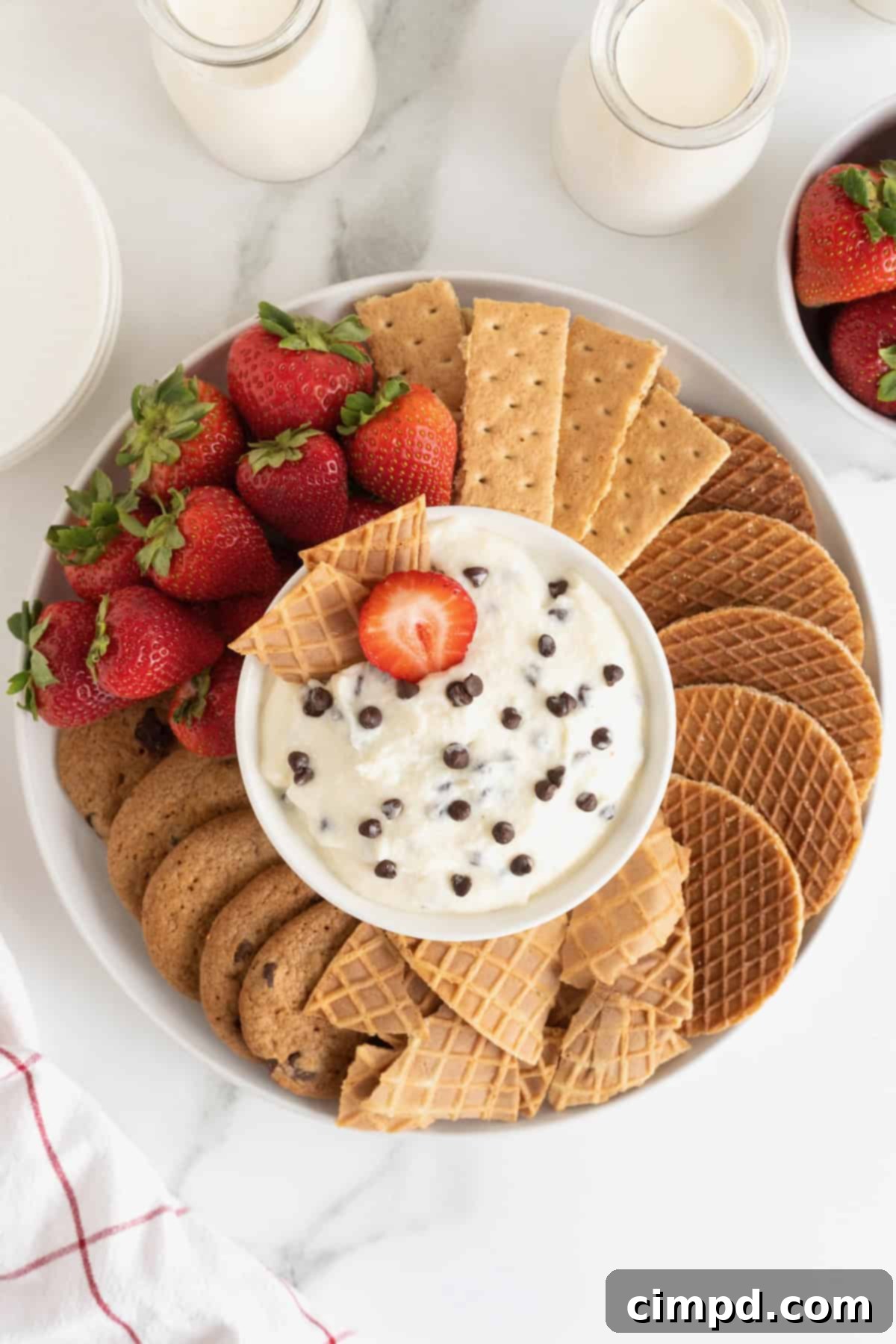A white serving platter filled with cookies, graham crackers, strawberries and sugar cone wafers. In the center of the dish is a white bowl filled with chocolate chip cannoli dip.